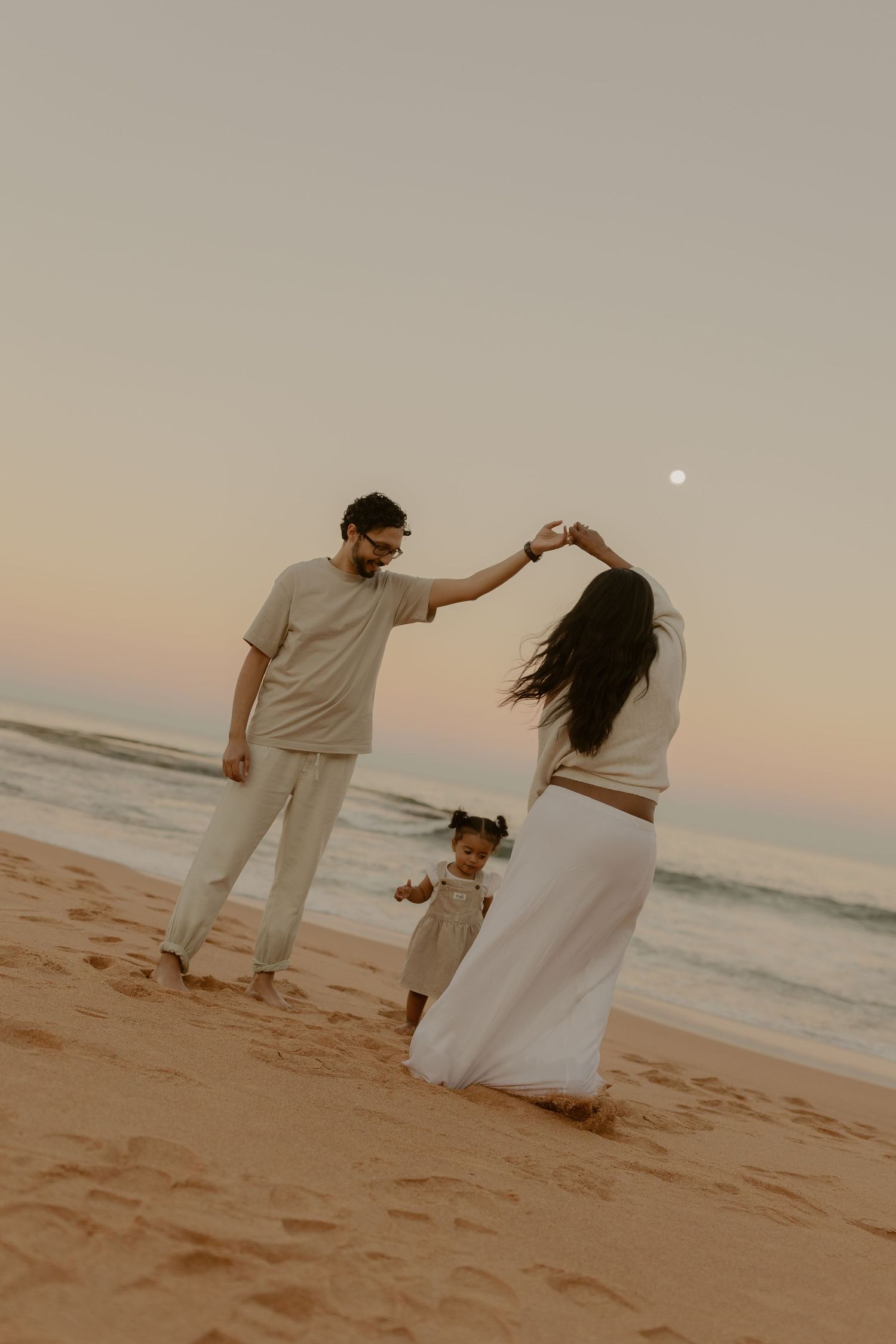 Family dancing on beach at sunset with toddler, parents holding hands overhead, ocean waves and moon in background.