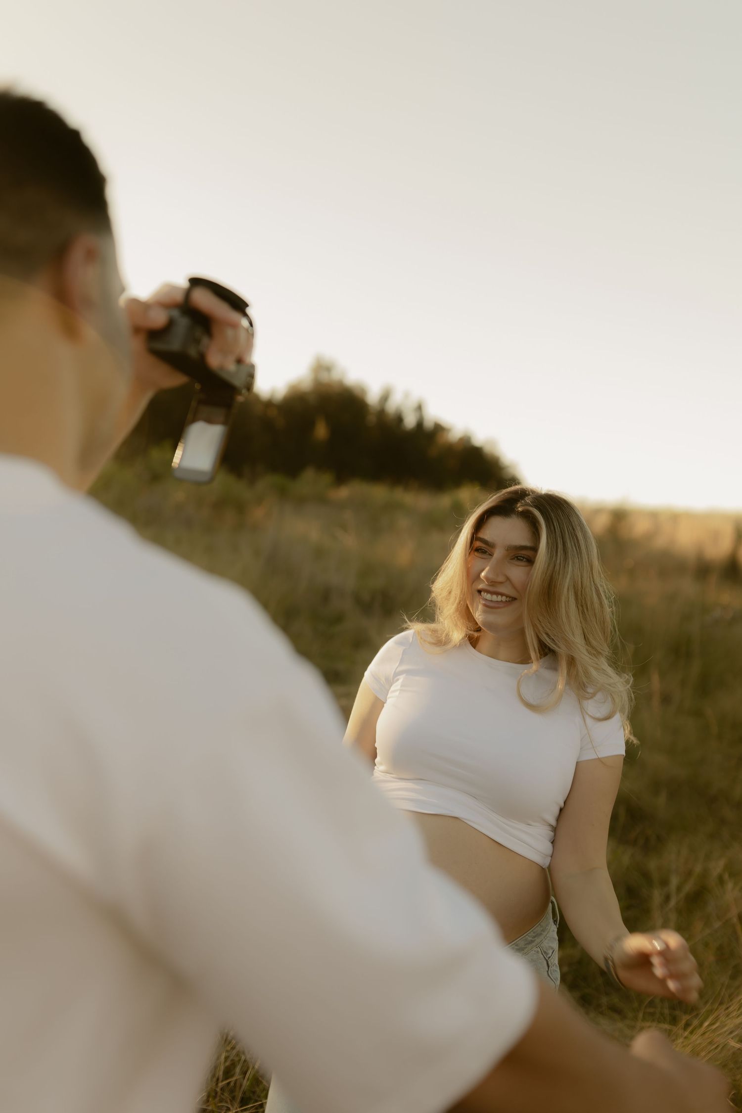 Man photographing smiling pregnant woman sitting in a sunny field during golden hour outdoors.