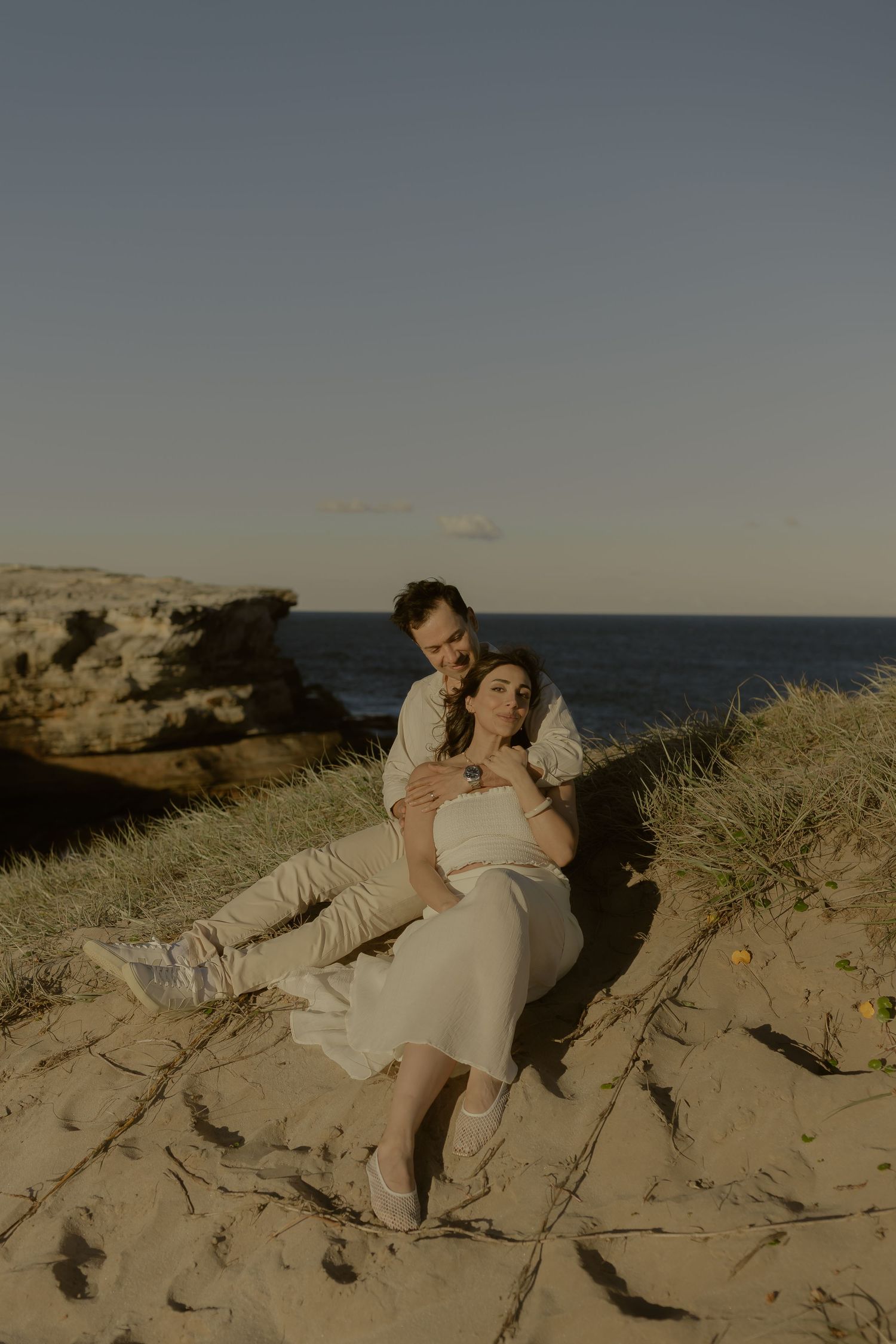 Couple in white wedding attire embracing on sandy dunes with ocean and rocky cliffs in background.