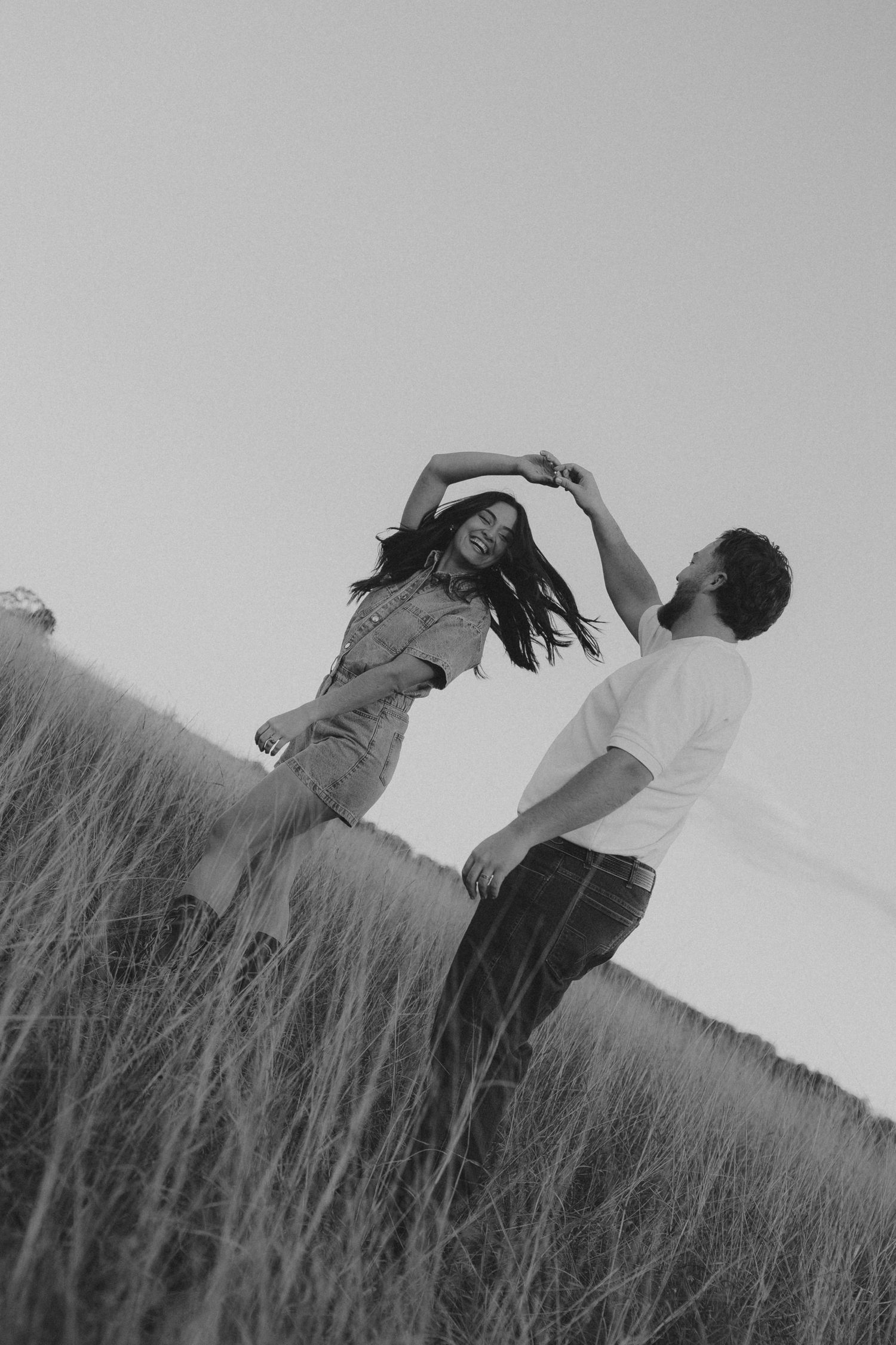 Black and white photo of a couple dancing joyfully in a grassy field, woman spinning with hair flowing.