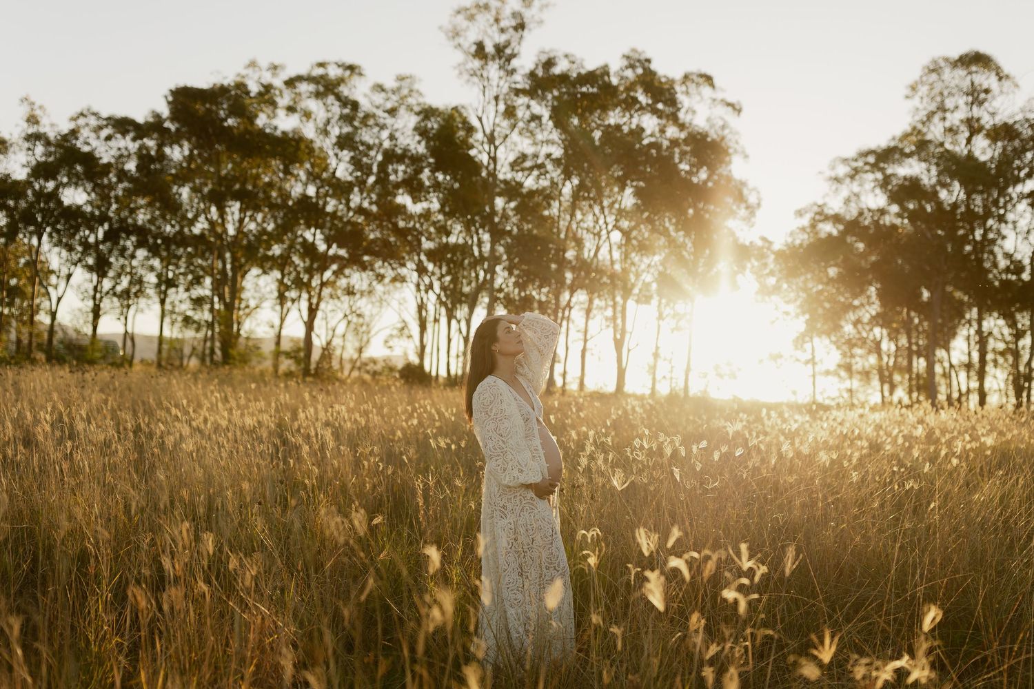 Woman in white dress standing in golden field at sunset with tall trees glowing in warm sunlight behind her.