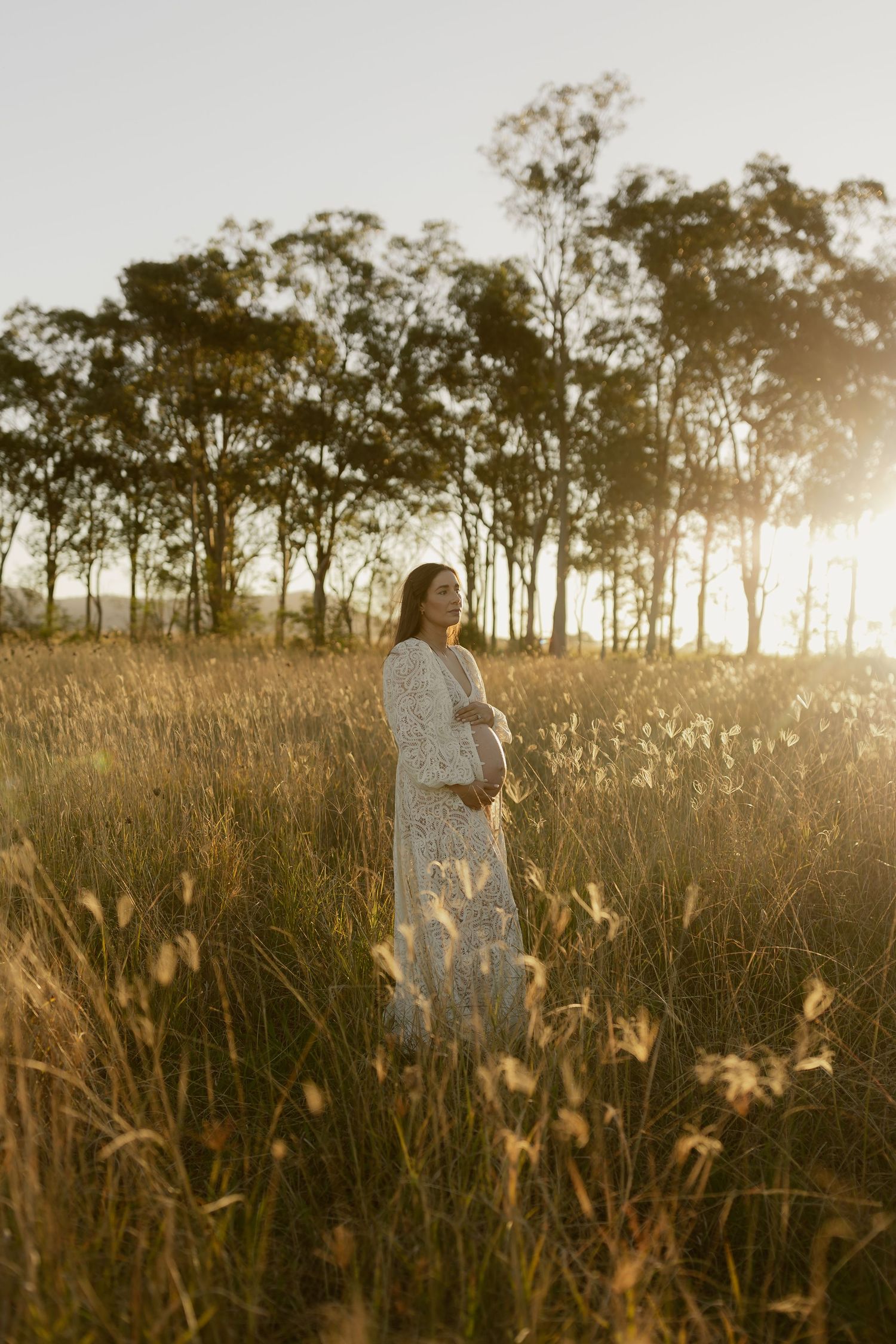 Pregnant woman in white floral dress standing in golden field at sunset with tall trees in background.