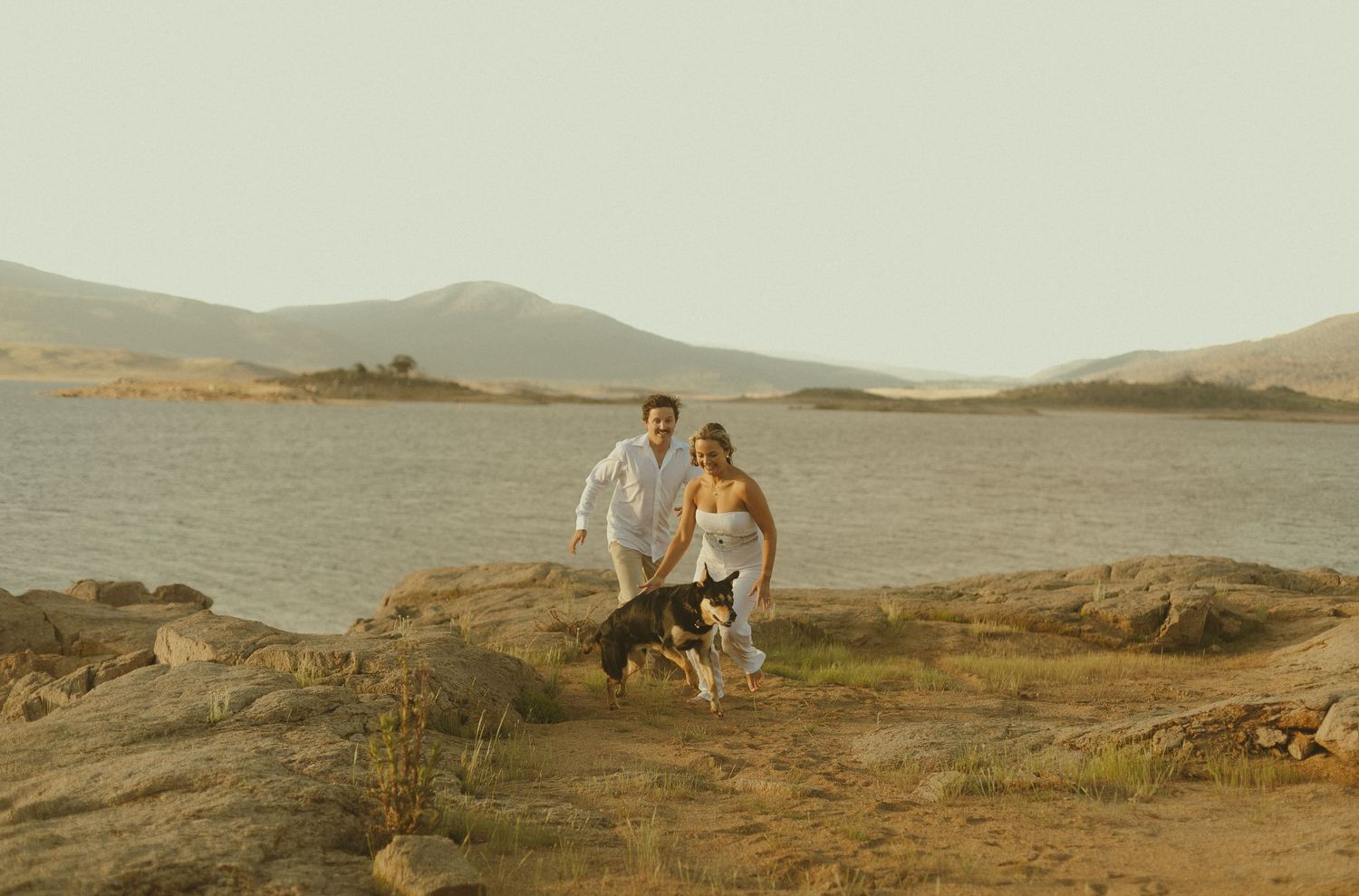 Two people run with their dog along rocky coastal terrain at sunset with mountains and water in the background.