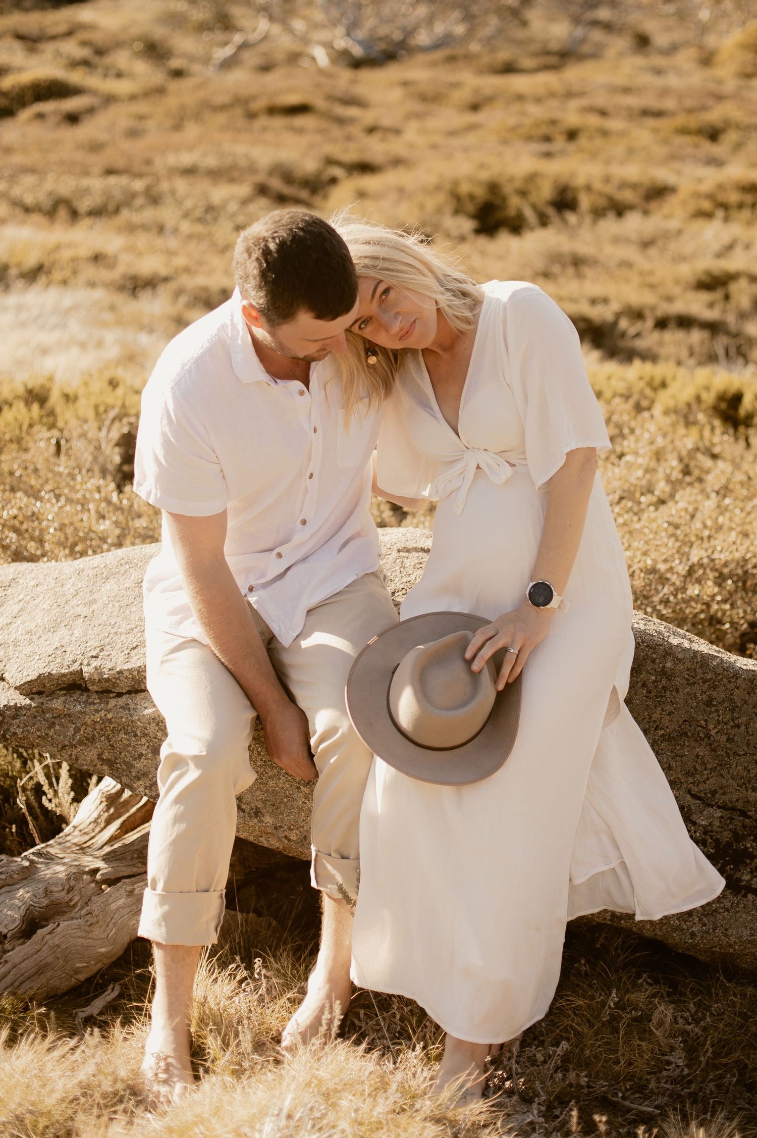 A romantic couple in white clothing sits together on a rock at sunset in a golden field holding a brown hat.