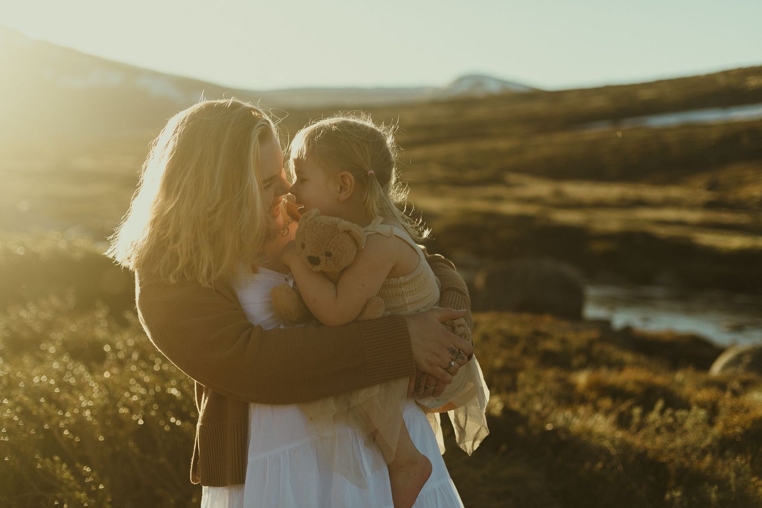 A tender family moment captured in golden sunlight with mountains and a river in the scenic background.