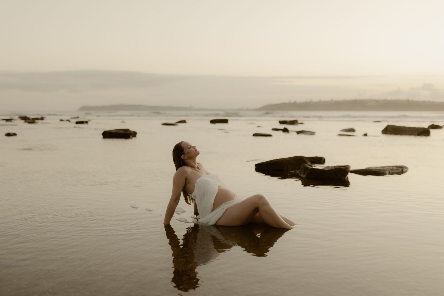An ethereal seascape photo with a figure posed among rocks in shallow water during golden hour.
