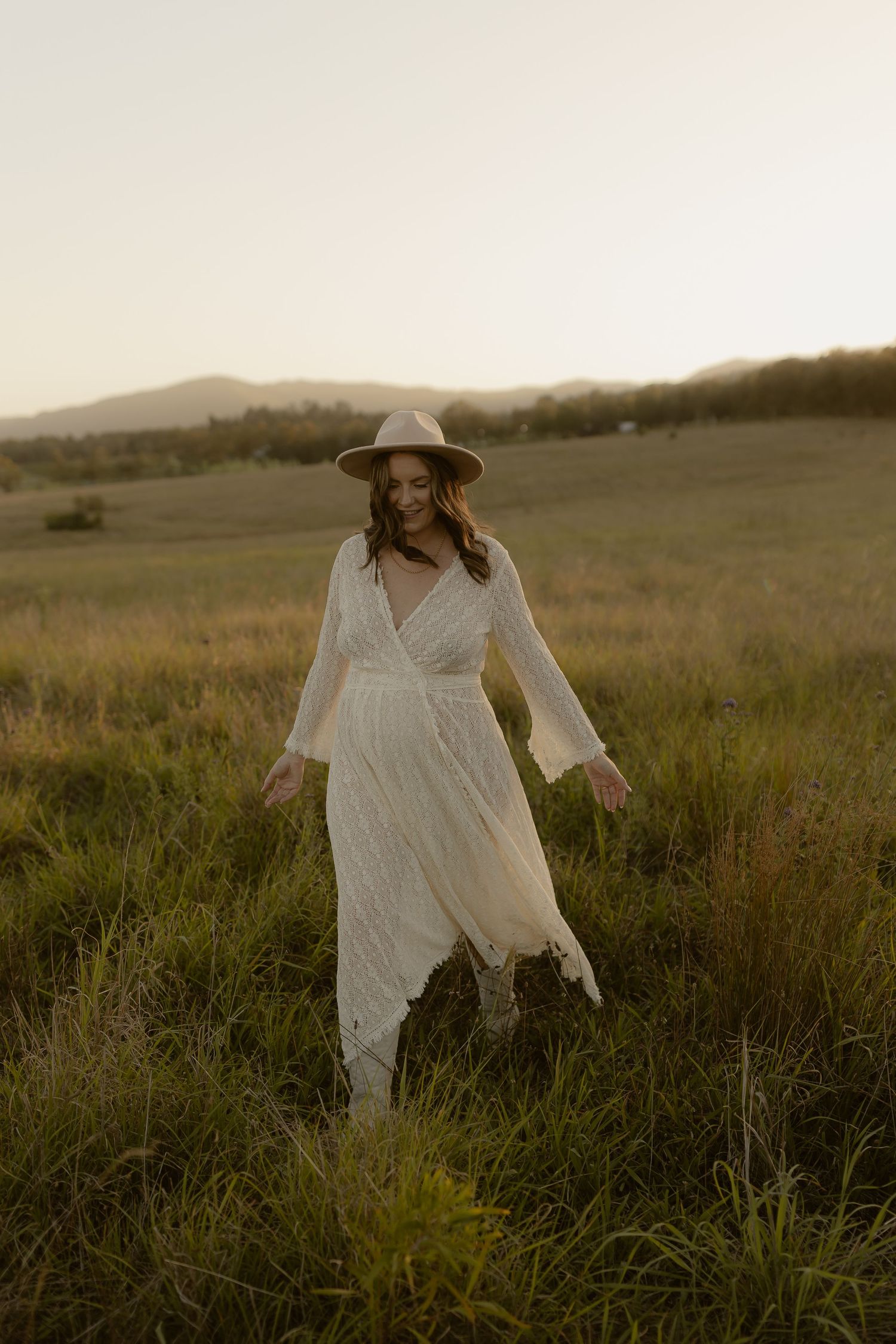 A person in a white dress and hat walks through a serene meadow at sunset.
