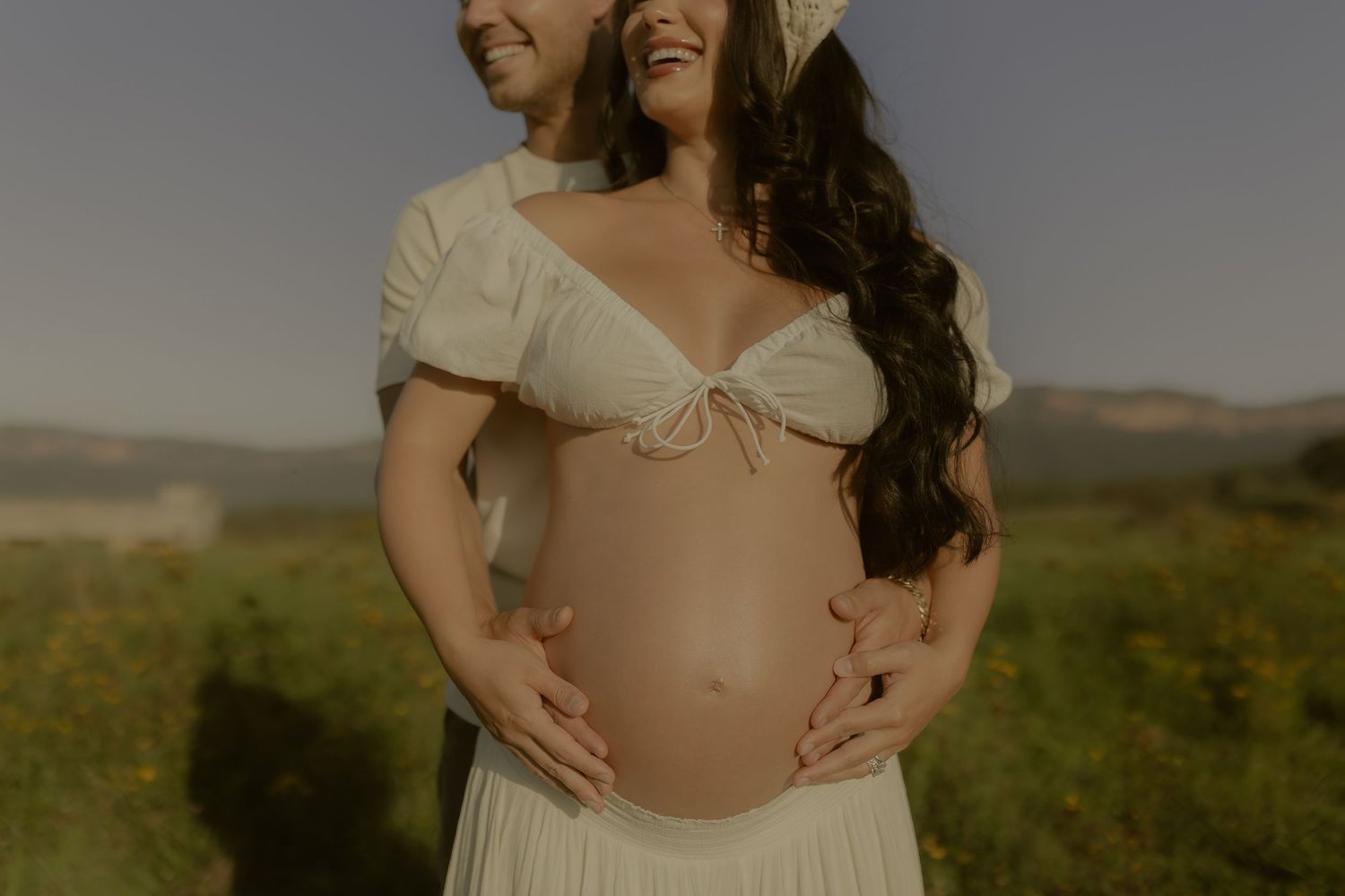 Romantic couple shares an intimate moment in a field with mountains and golden sunlight.