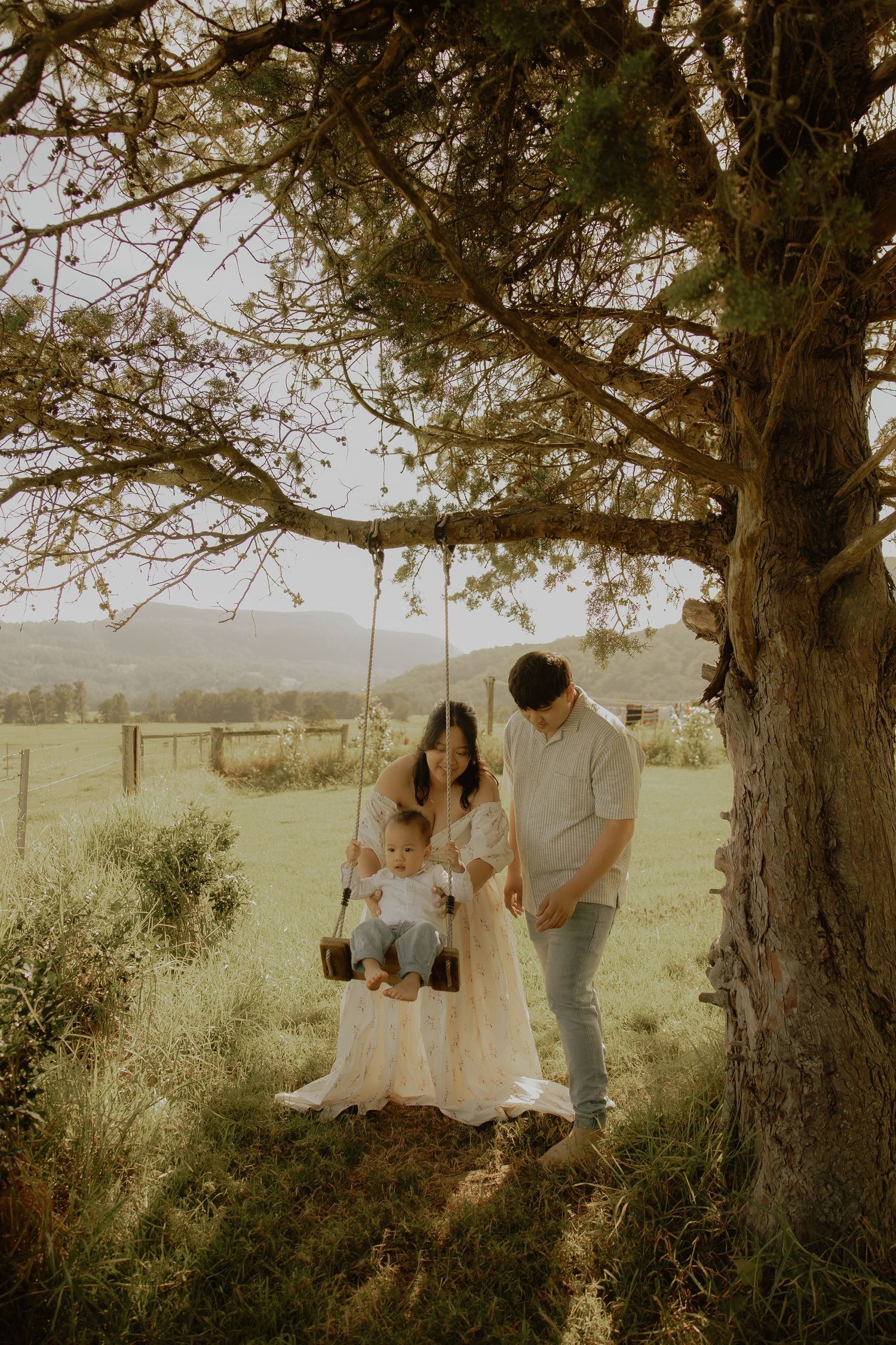Family with baby on tree swing in lush green field, mother in white dress, golden hour light.