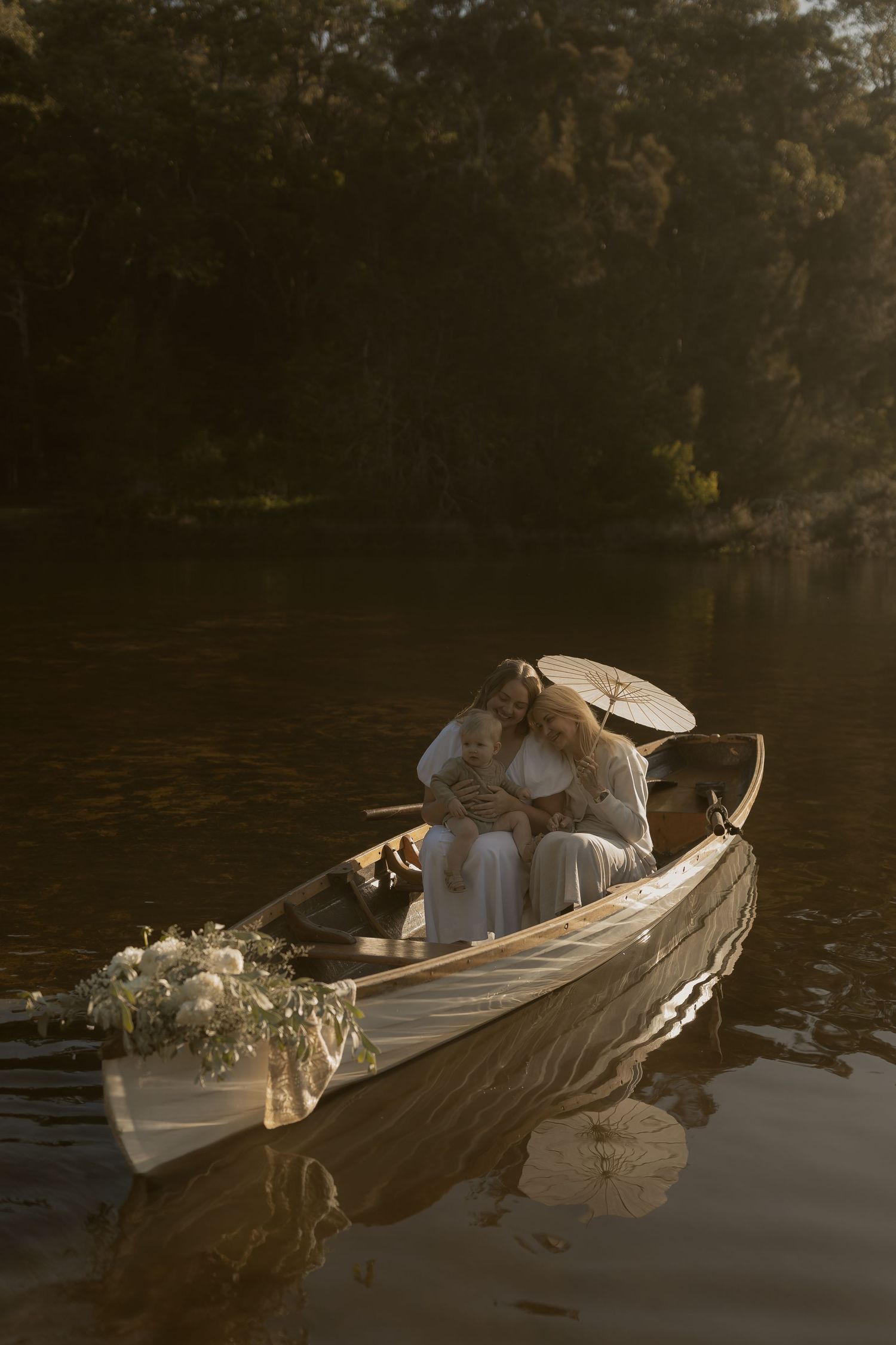 Couple embracing in a white wooden canoe on a calm lake, surrounded by lush trees in golden hour light.