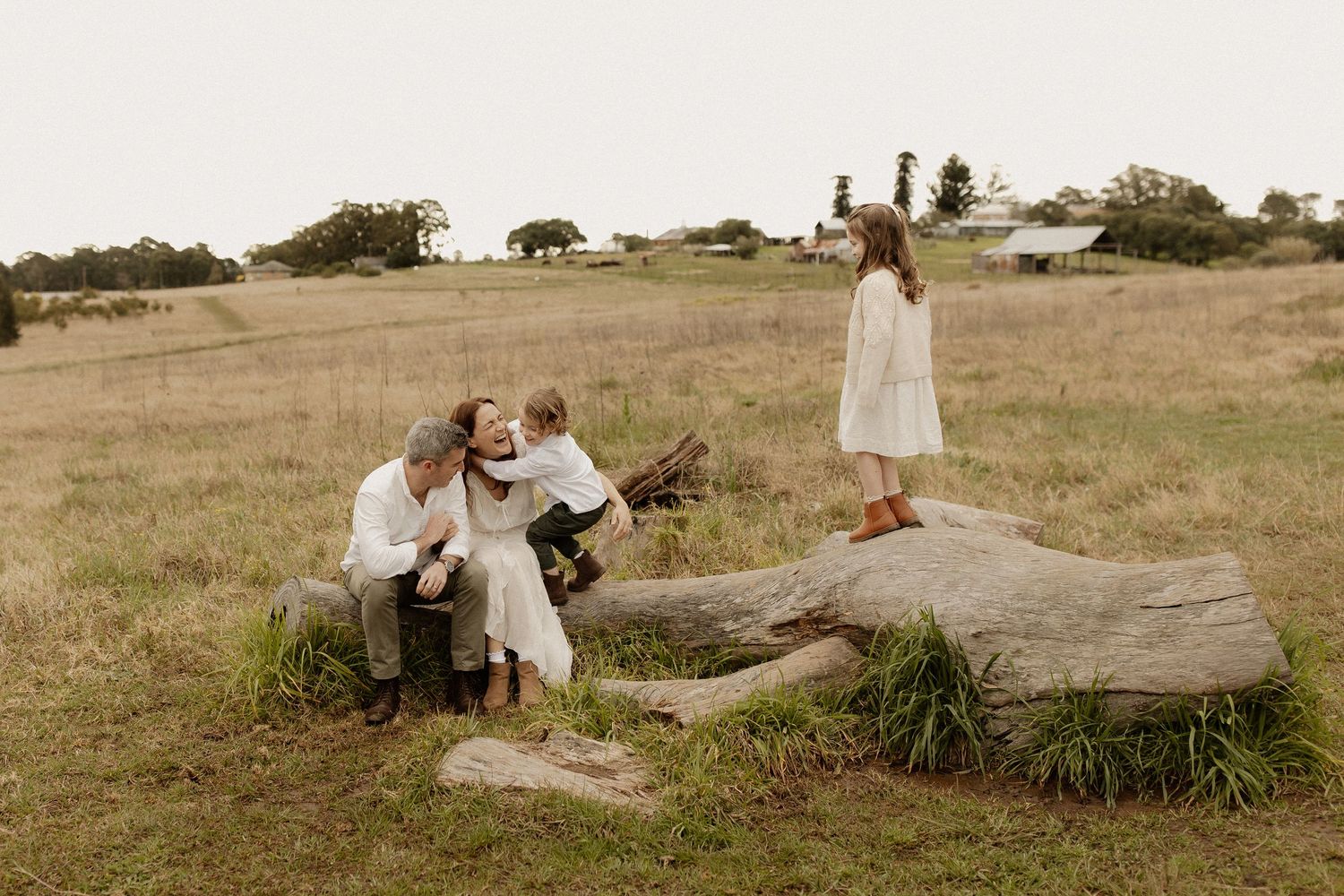 People gather around fallen tree trunk in rustic outdoor setting with dried grass field and trees in background.