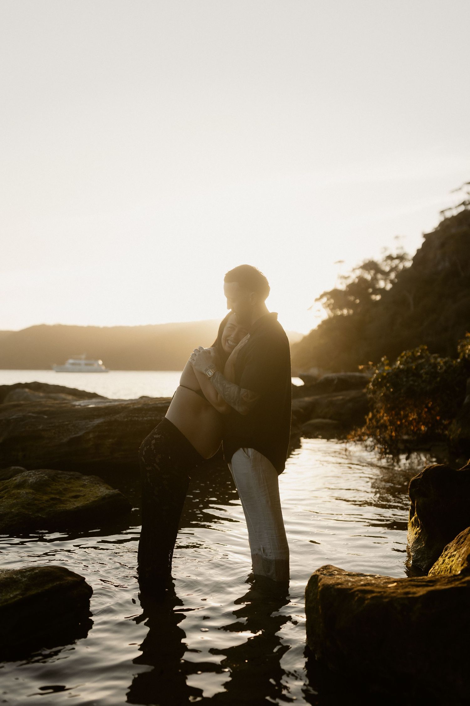 Silhouettes of a couple embracing on rocky coastline during golden sunset by the ocean.