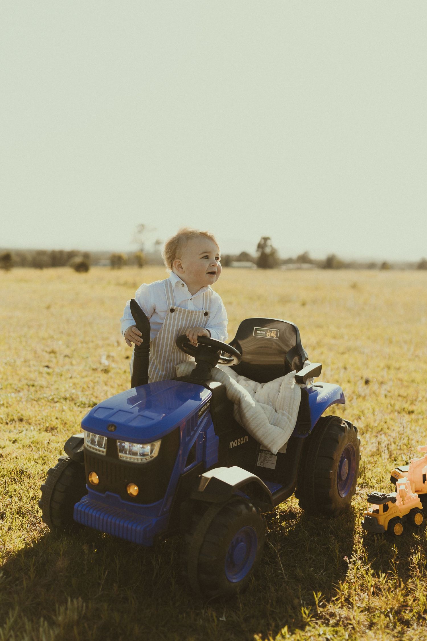 A toddler rides a blue toy tractor in a sunny field.