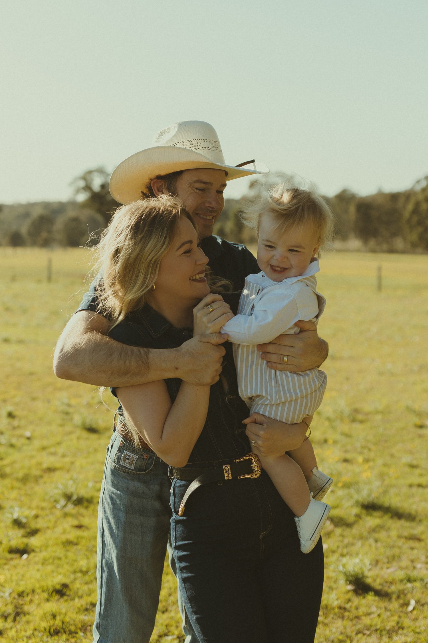 Family members embrace while wearing cowboy hats in a sun-drenched field.