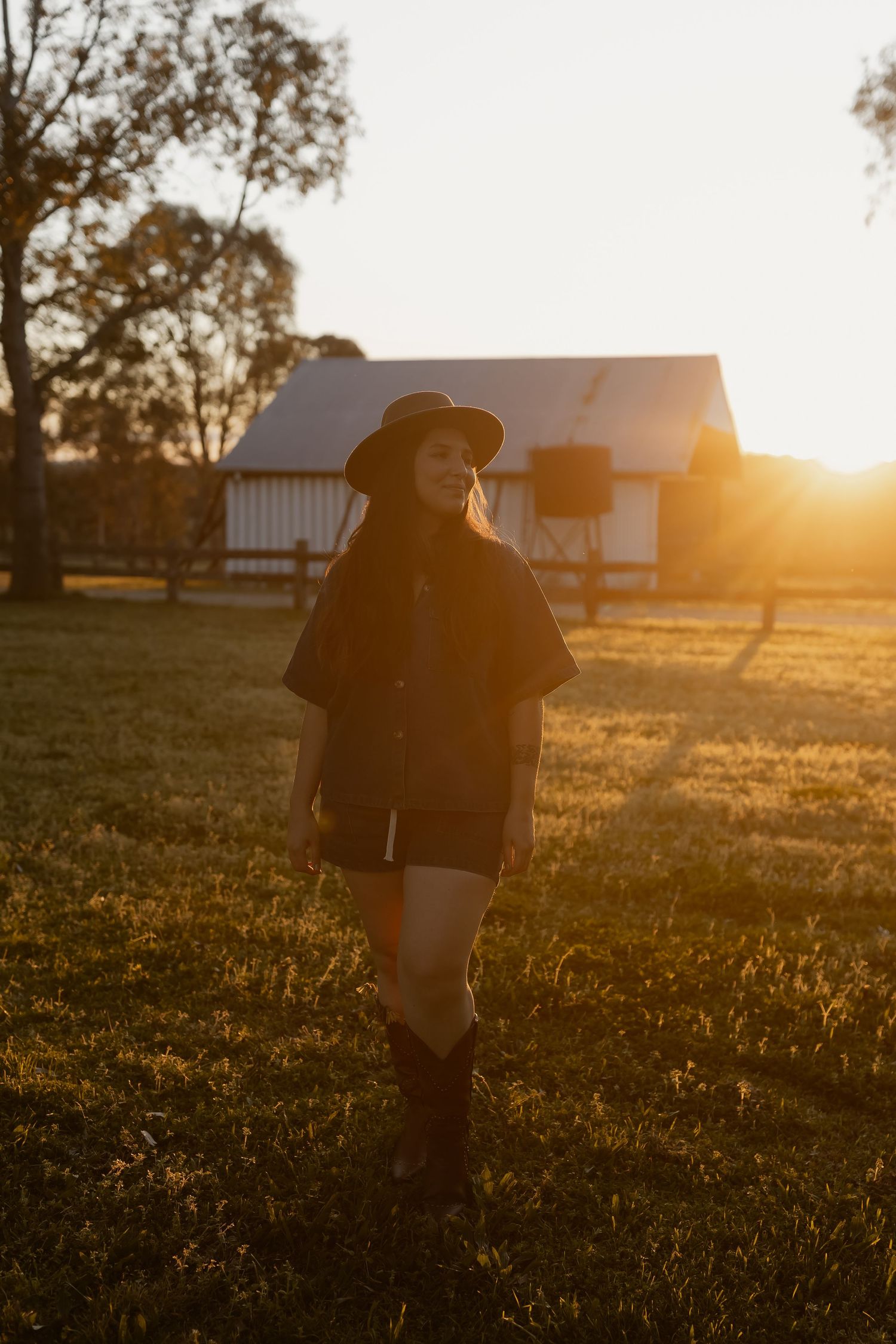 Silhouetted figure in cowboy hat walking through golden grass field at sunset with farm buildings in background.