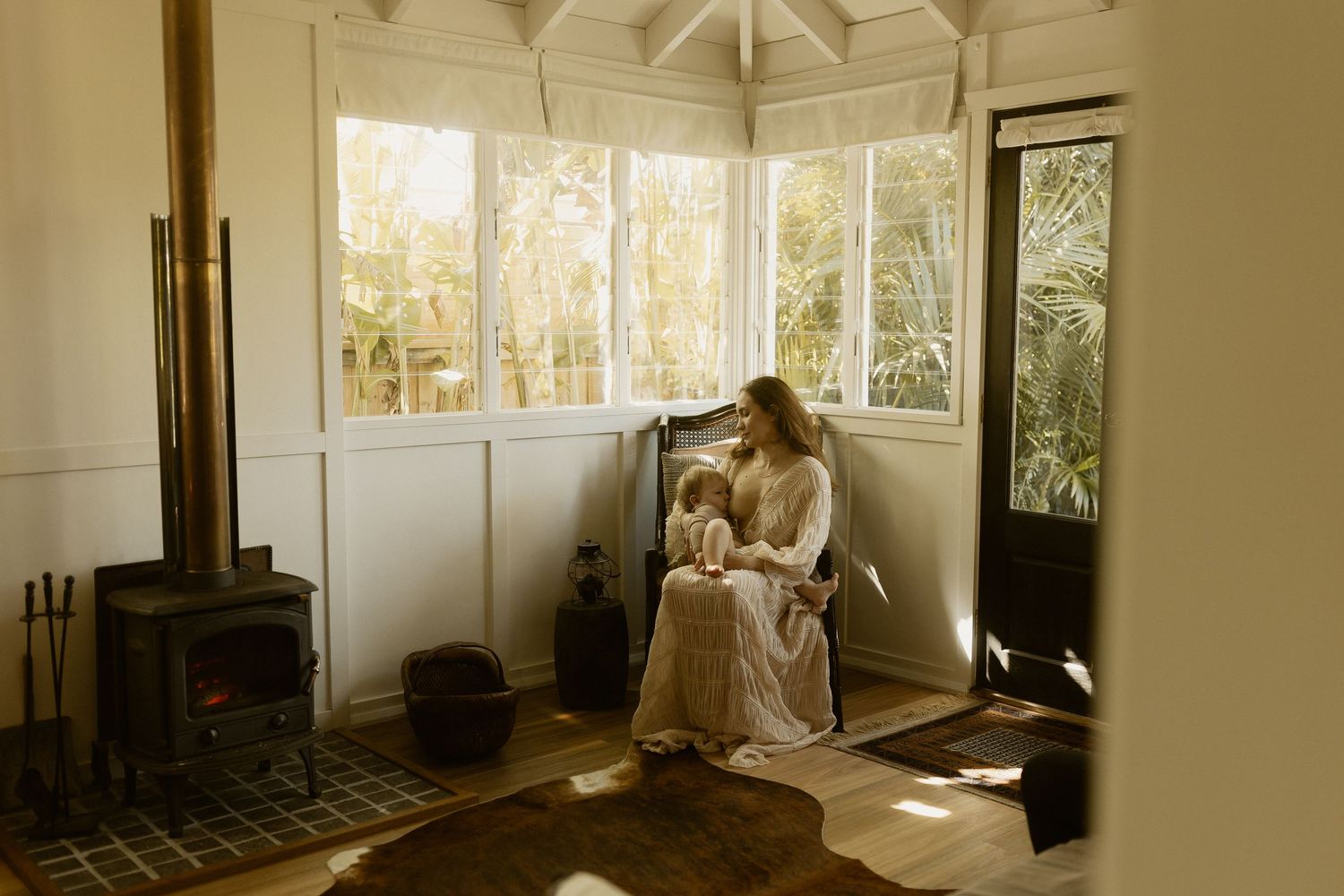 A cozy sunlit room with a wood stove features someone seated in a corner reading near large windows.