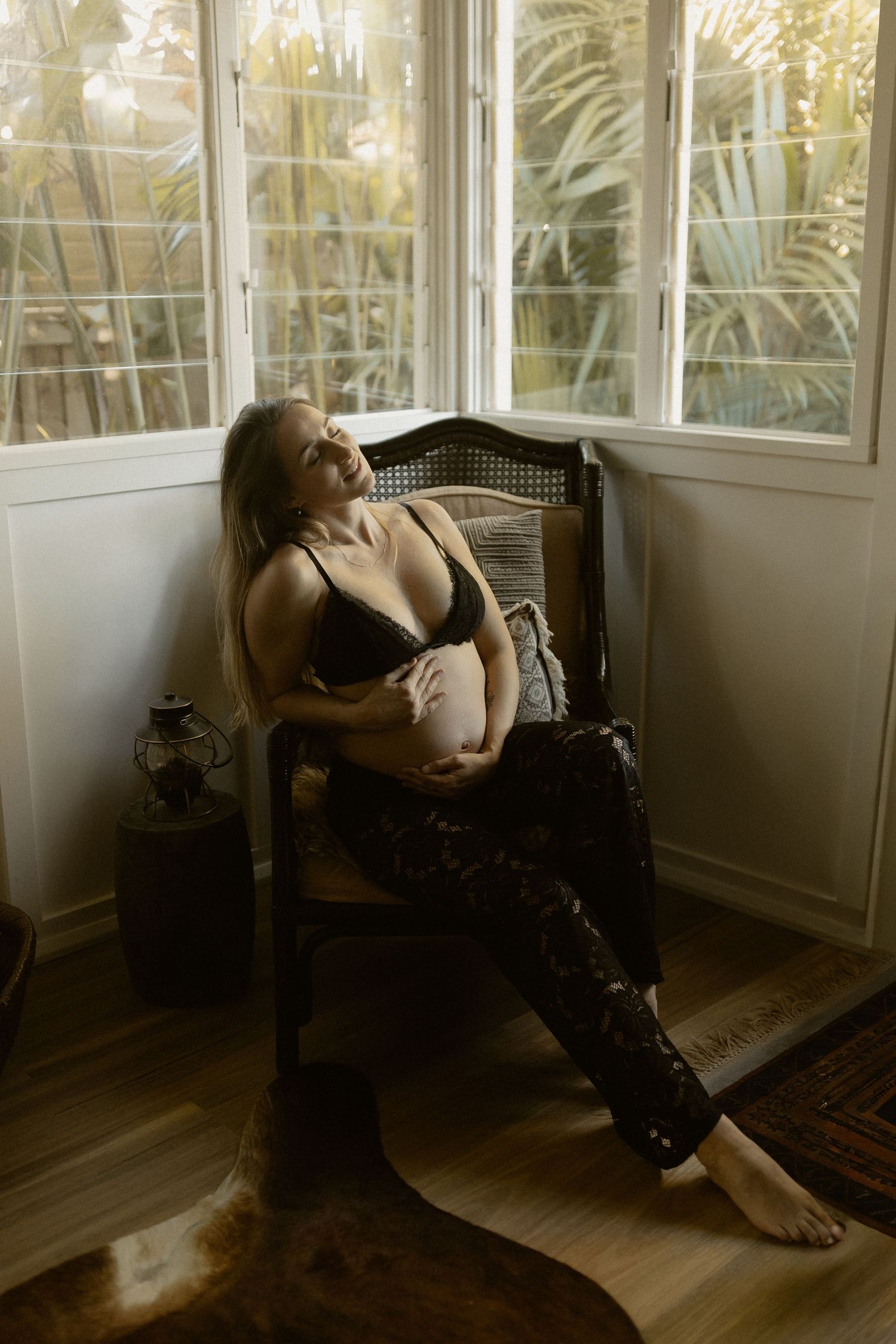 A moody boudoir photoshoot in a sunlit corner with antique chair and palm trees visible through windows.