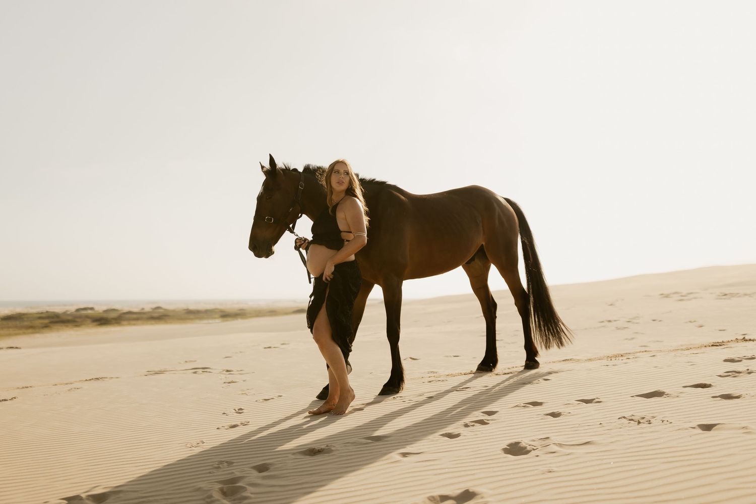 A romantic beach photoshoot featuring a horse and couple walking along the sandy shoreline at sunset.