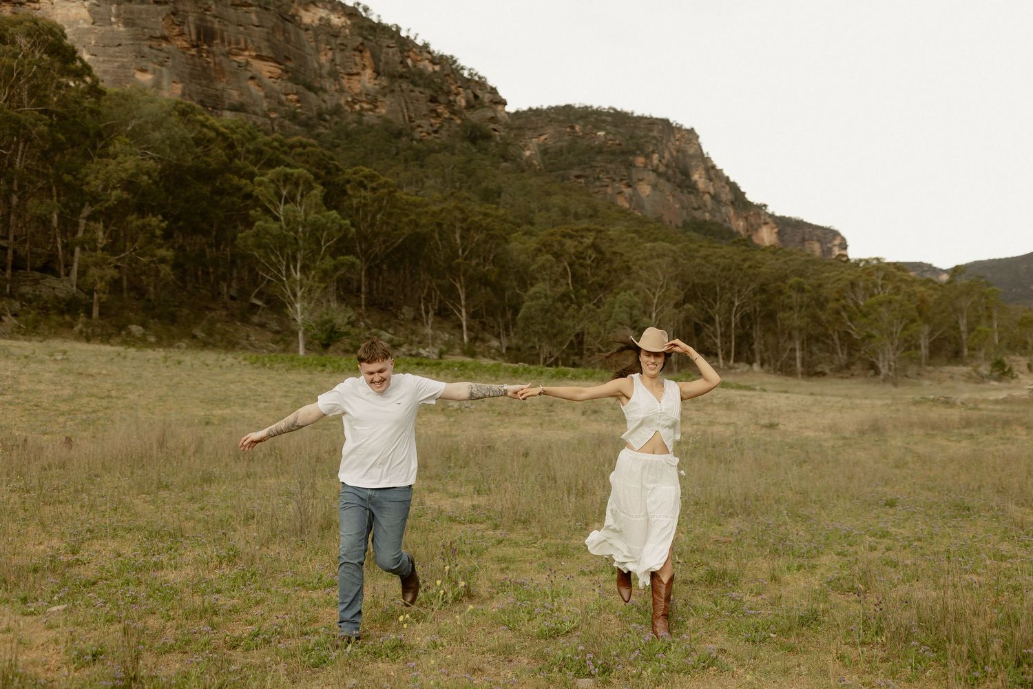 Two people run joyfully through a grassy meadow with mountains and trees in the background at sunset.