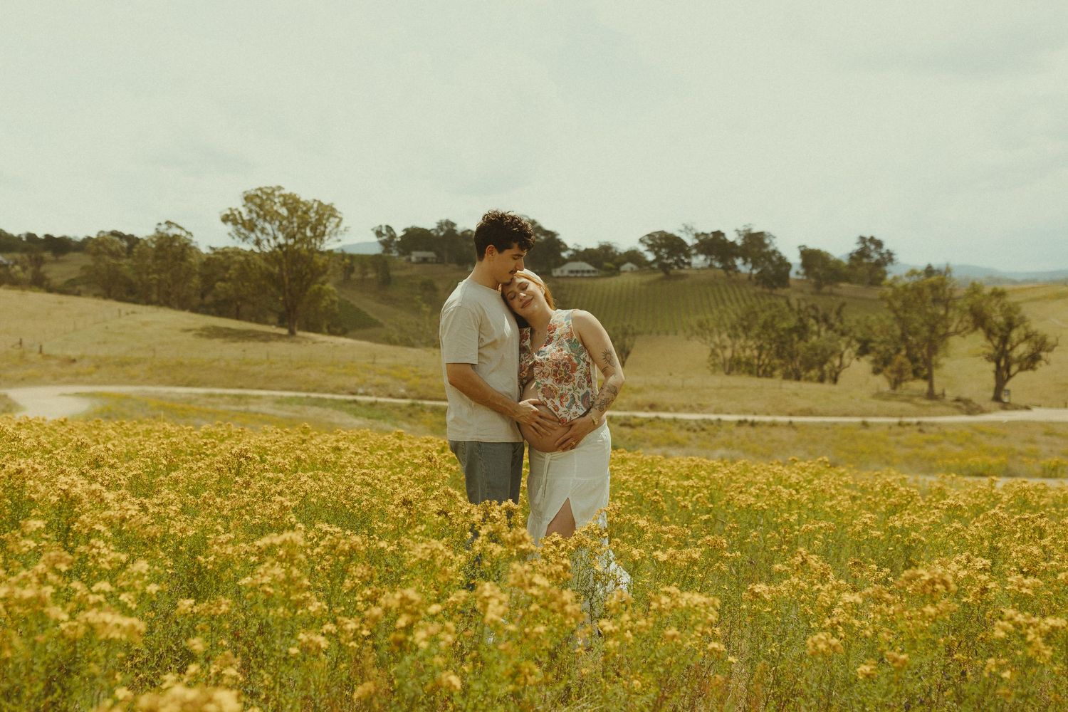 A romantic couple stands together in a field of vibrant yellow wildflowers with rolling hills in the background.