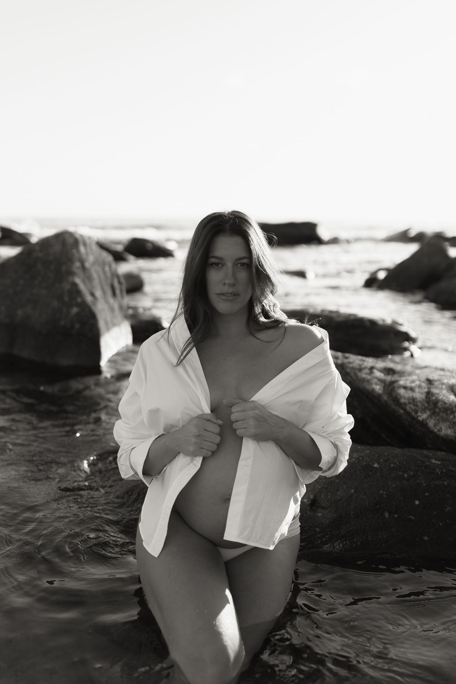 Woman in flowing white shirt poses artistically among beach rocks at sunset.