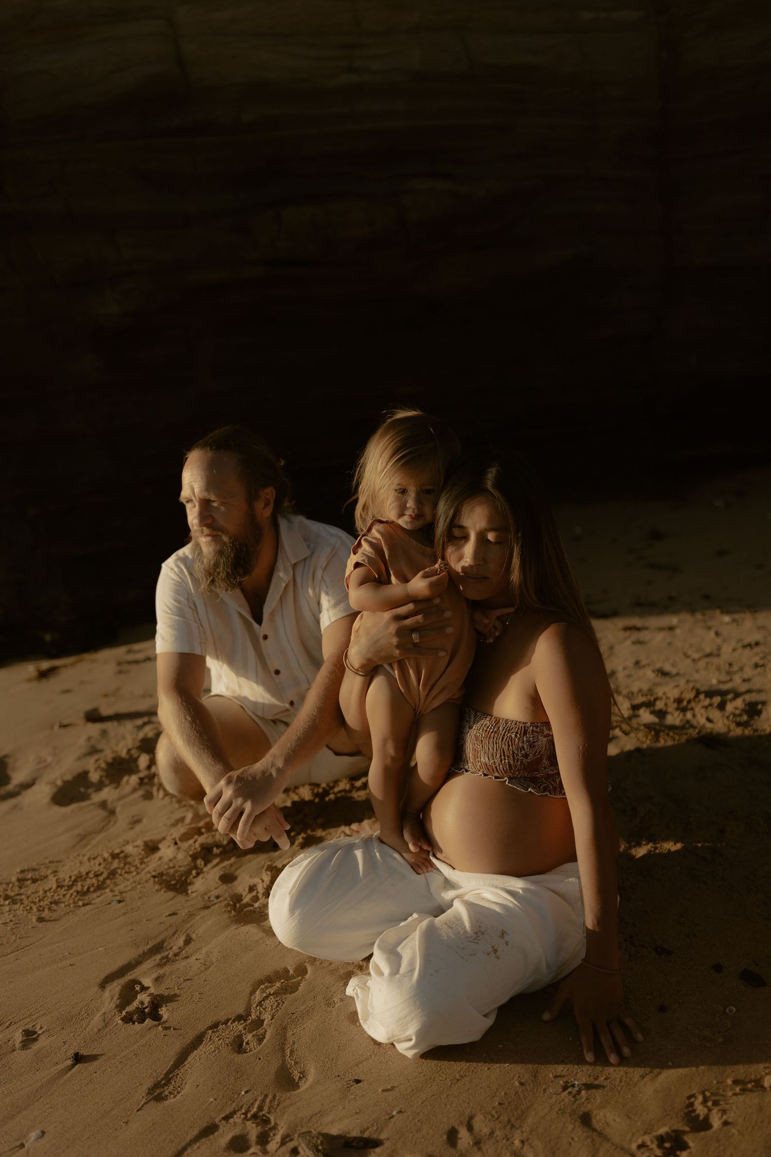 Family sits together on sandy beach at sunset creating intimate maternity portrait in warm golden light.