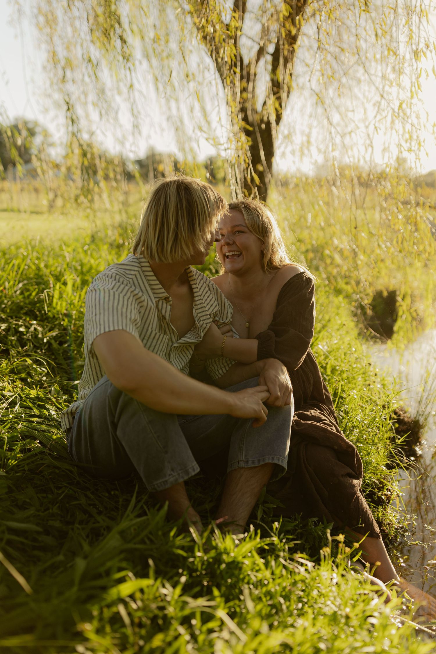Two people share a tender moment while sitting in tall grass at sunset with golden sunlight streaming through trees.