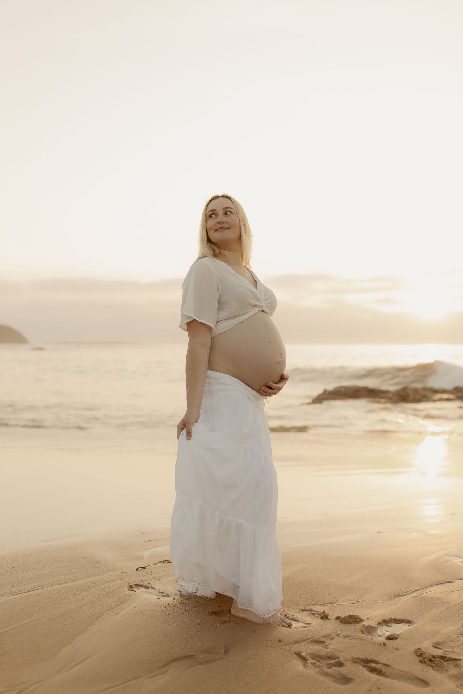 A serene maternity session captures moments on a beach at golden hour with a white flowing dress.