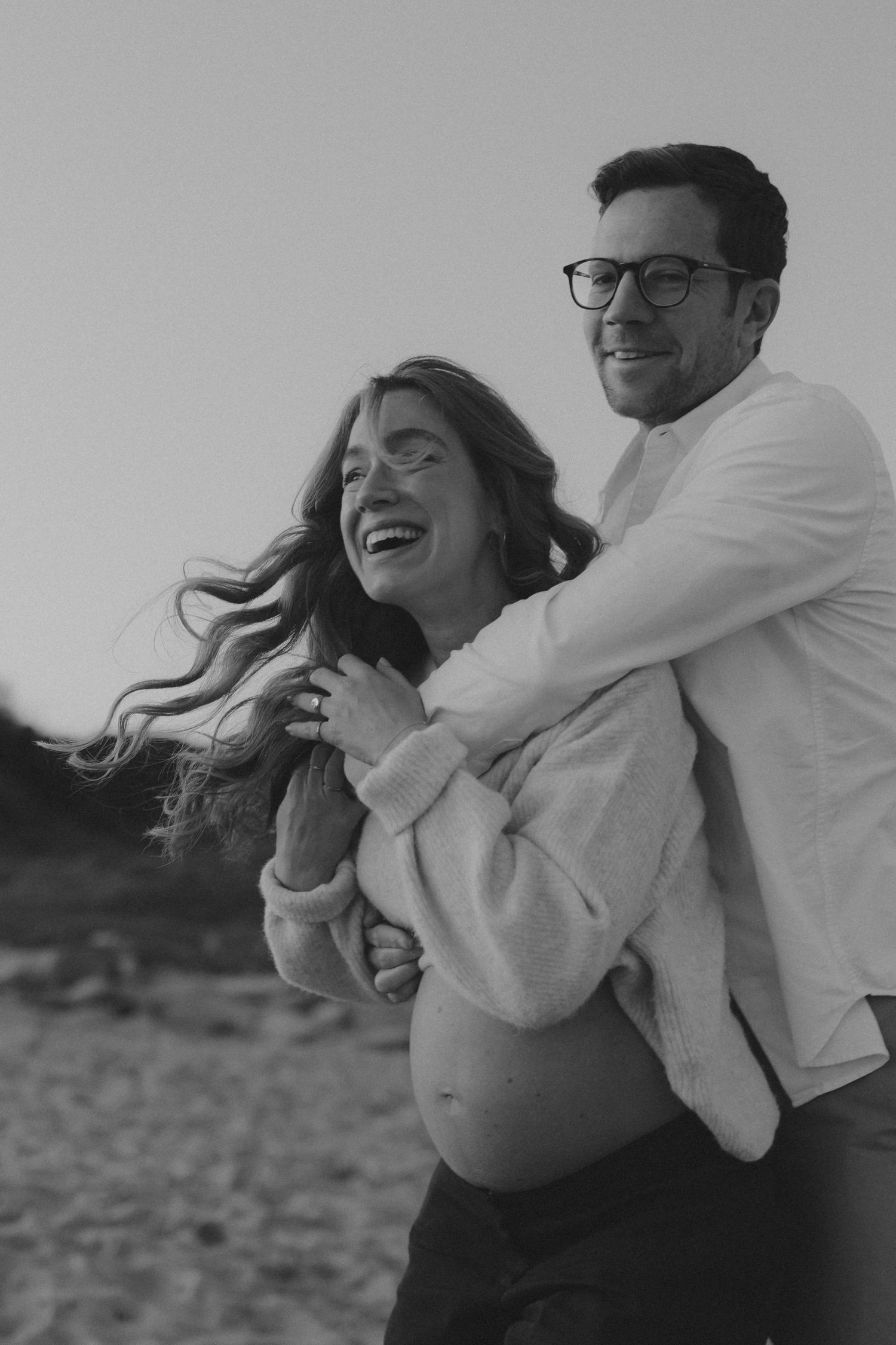 Black and white photo of a laughing pregnant woman embraced by a smiling man wearing glasses on a beach.