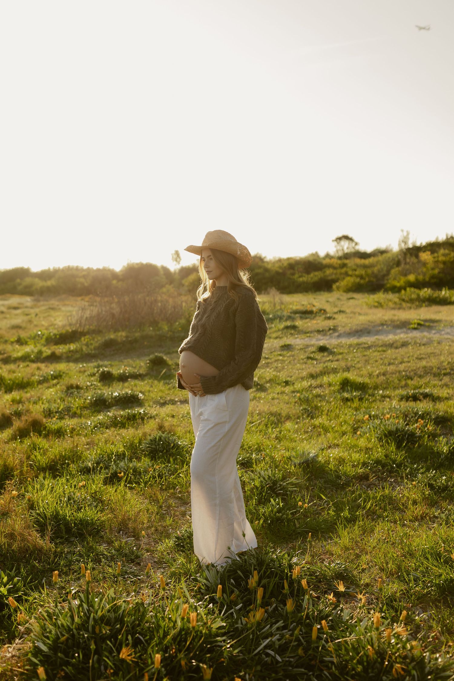 Pregnant woman in hat and white skirt stands in sunlit green field, cradling her baby bump at golden hour.