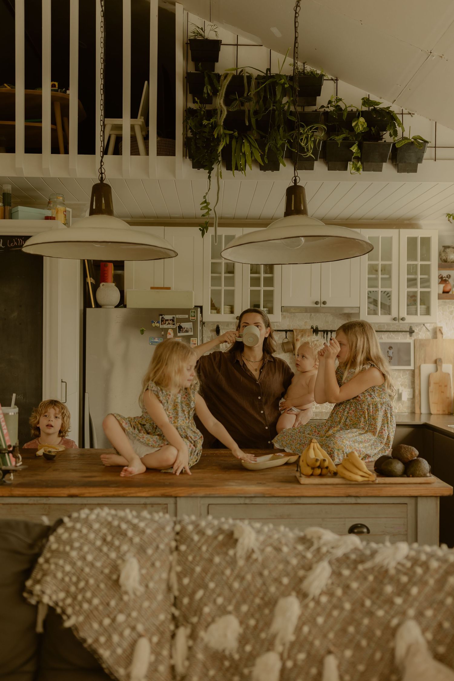 Mother and four children laughing together at a wooden kitchen island with hanging plants above.
