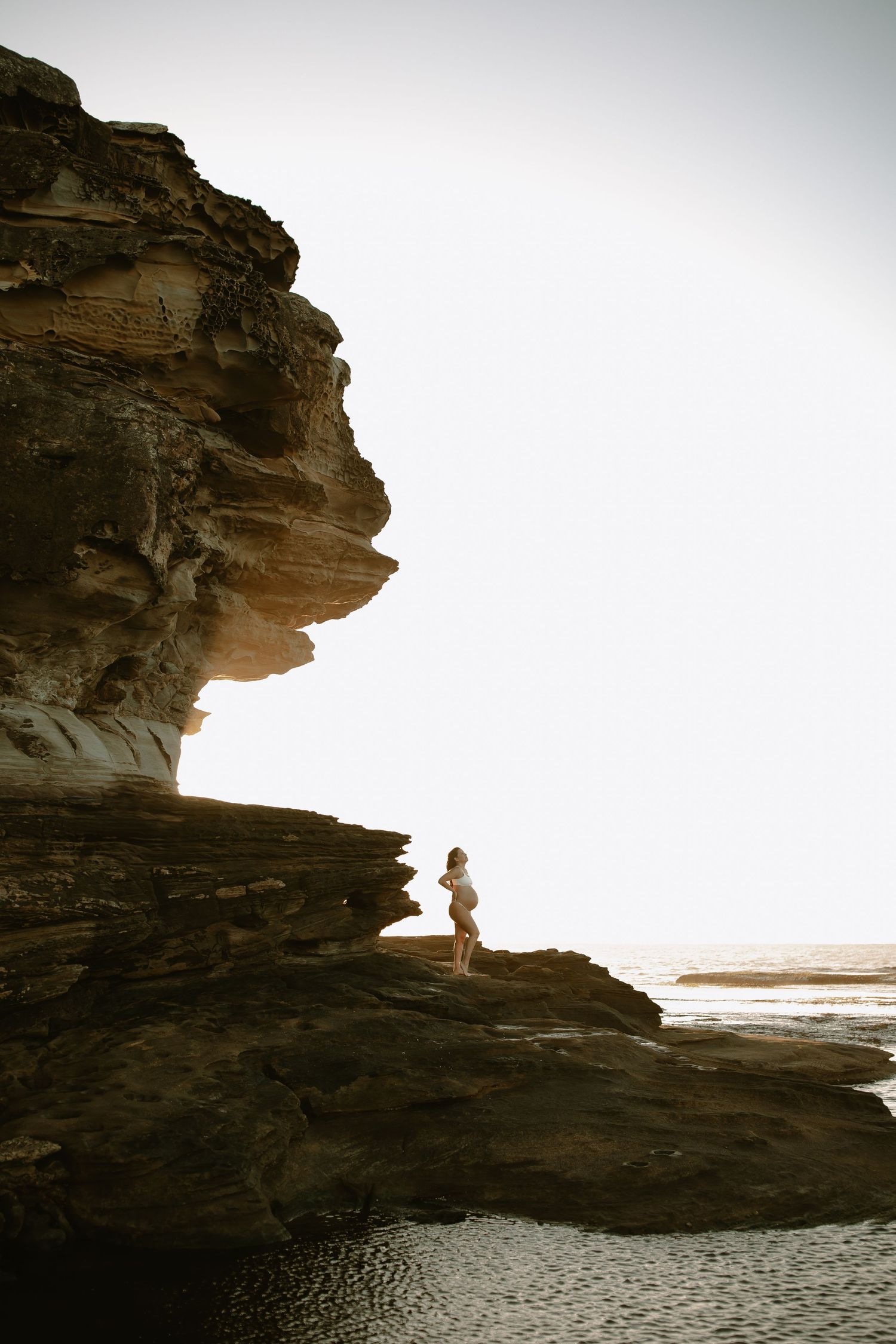 Silhouette of a small figure standing on dramatic coastal cliffs against a bright sky at sunset.