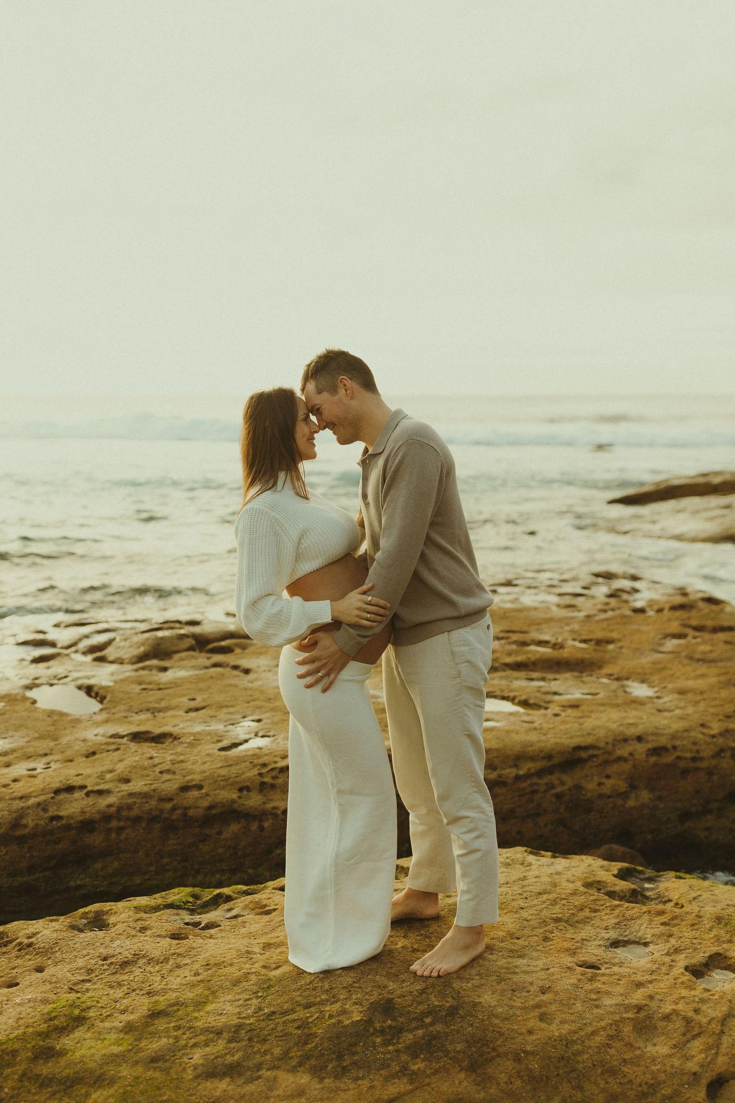 A couple in white outfits sharing an intimate moment on coastal rocks during golden hour at the beach.
