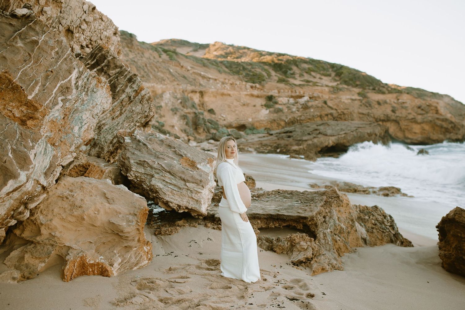 Figure in flowing white dress stands among rugged coastal cliffs and beach landscape as waves crash on rocky shore.