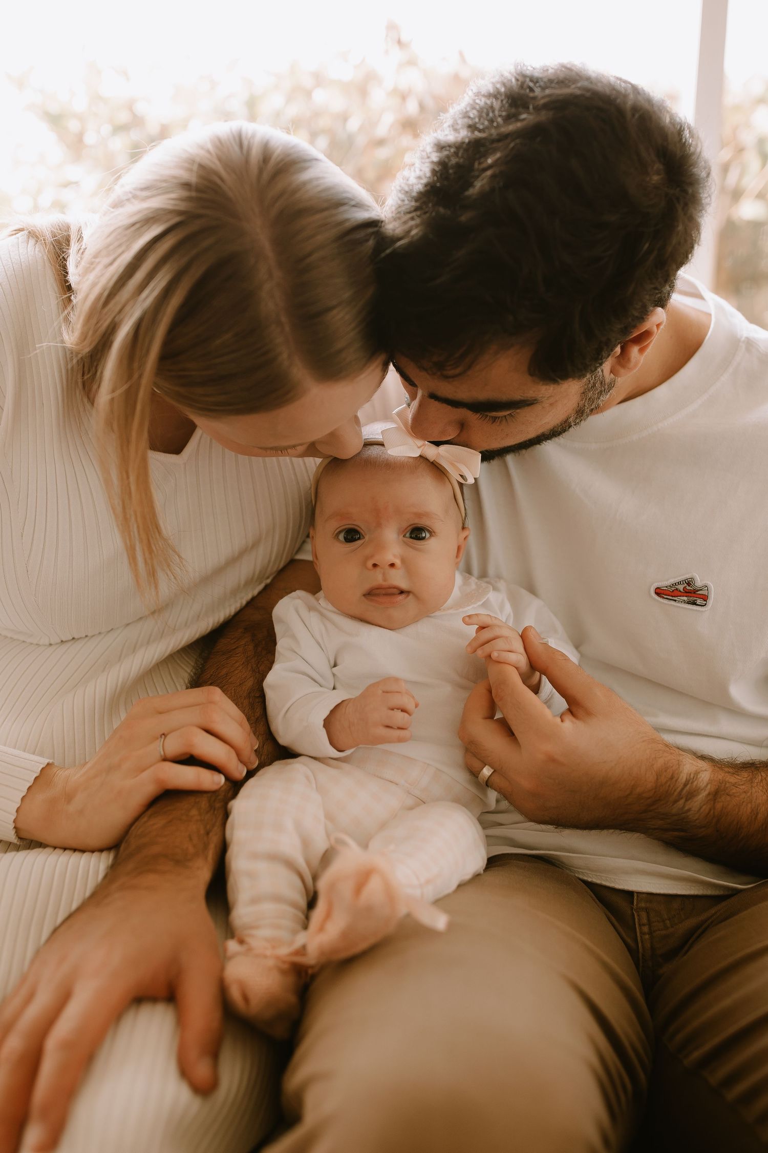 Parents lean in close to their infant baby wearing white who is lying on their lap in warm golden lighting.