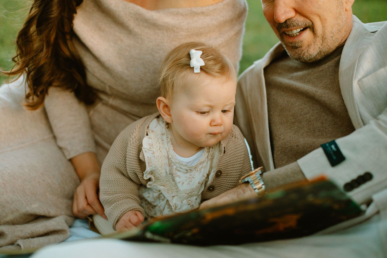 Parents and baby looking at picture book together while sitting on picnic blanket outdoors.