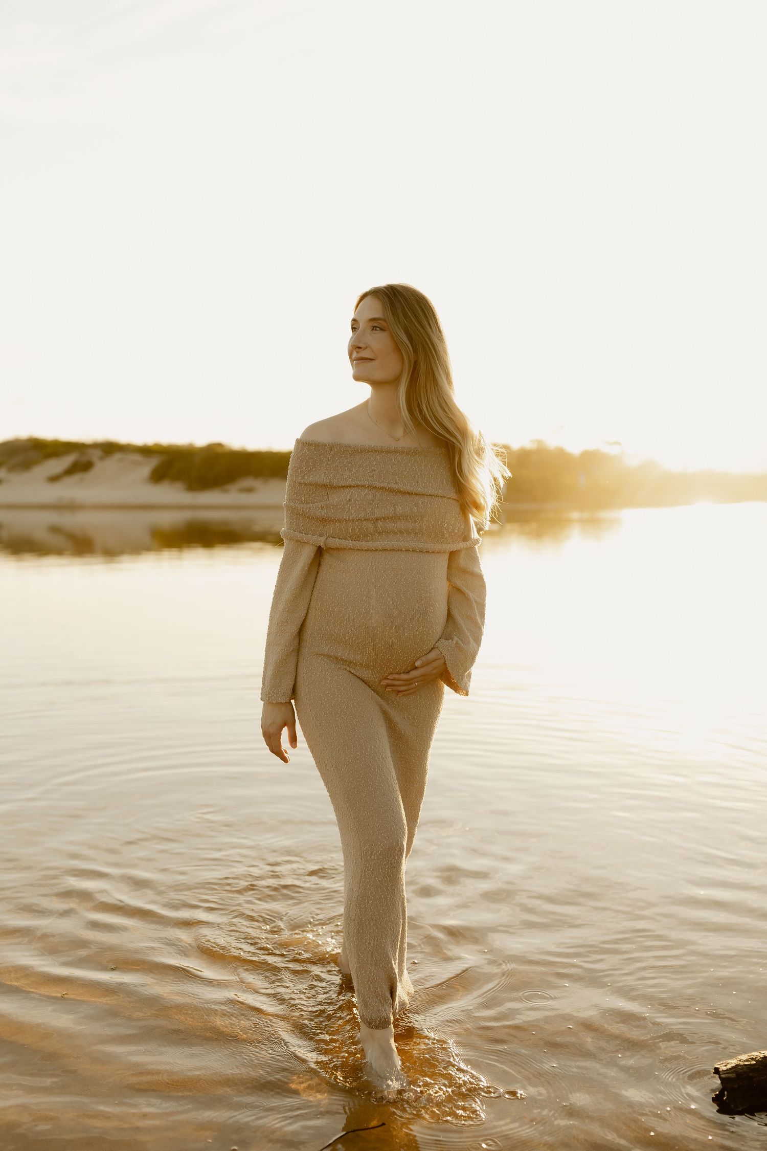 A person in white clothing walks through shallow water at sunset creating ripples on the calm surface.
