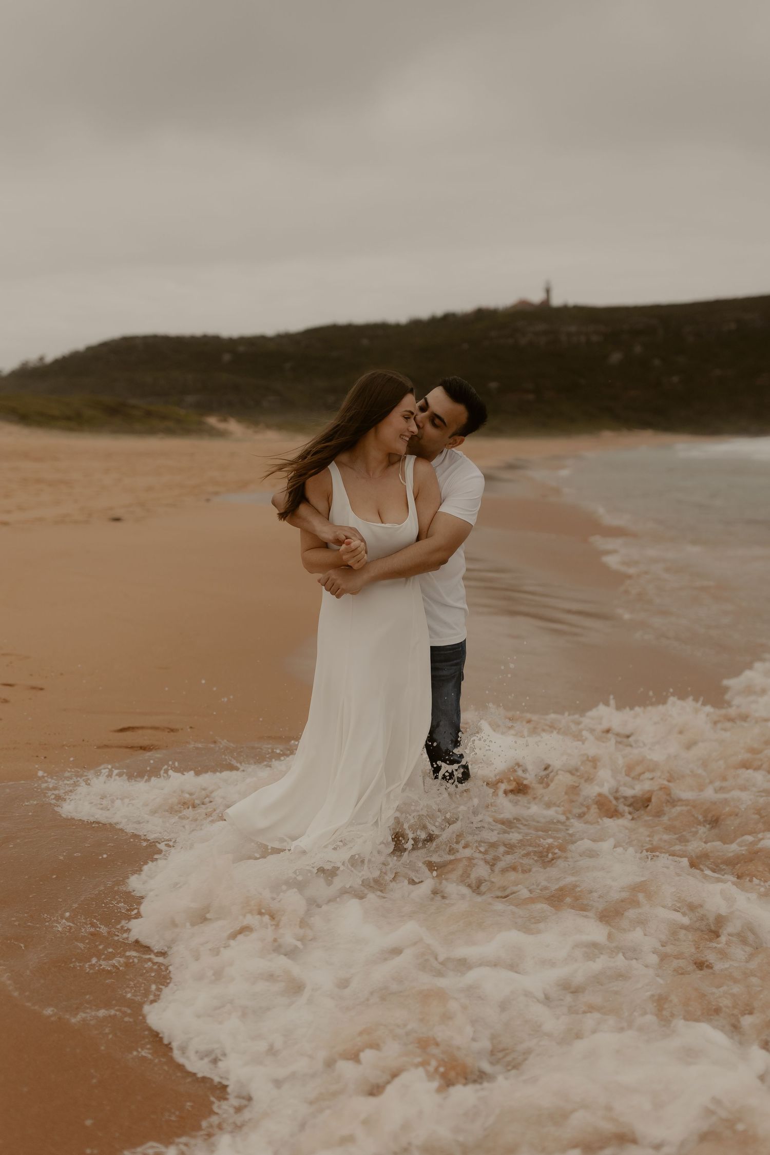 A romantic couple stands in the surf as waves crash around them during sunset at the beach.