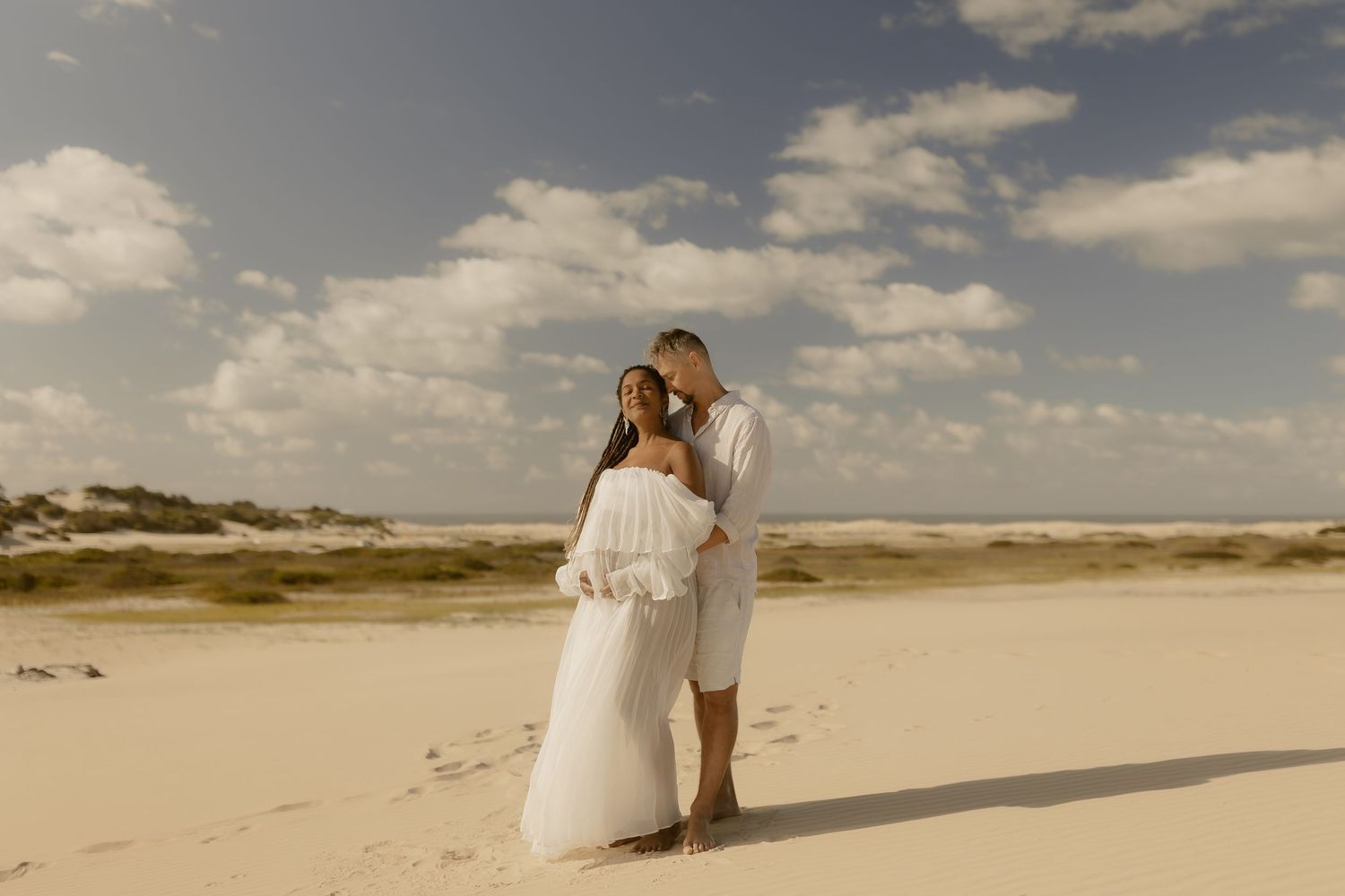 Romantic beach wedding couple embraces on sandy shore with dramatic cloudy sky at sunset.