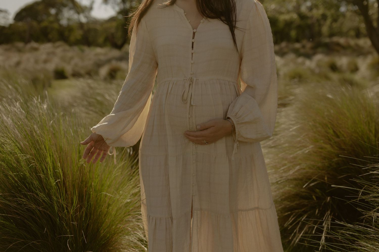 A person in a flowing white dress poses in a sunny field surrounded by tall ornamental grasses.