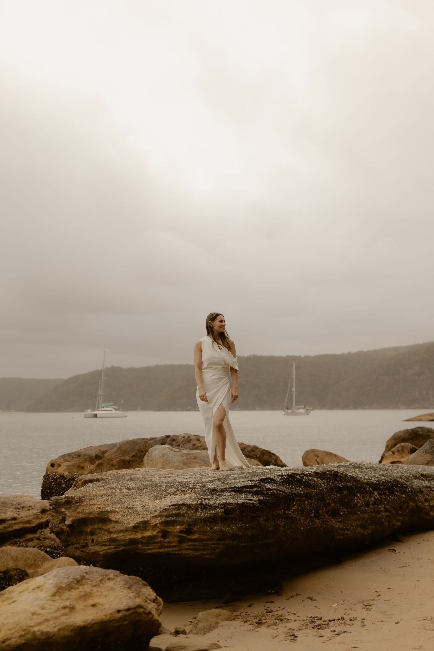 A couple in white attire stands on coastal rocks overlooking a misty harbor with boats in the distance.