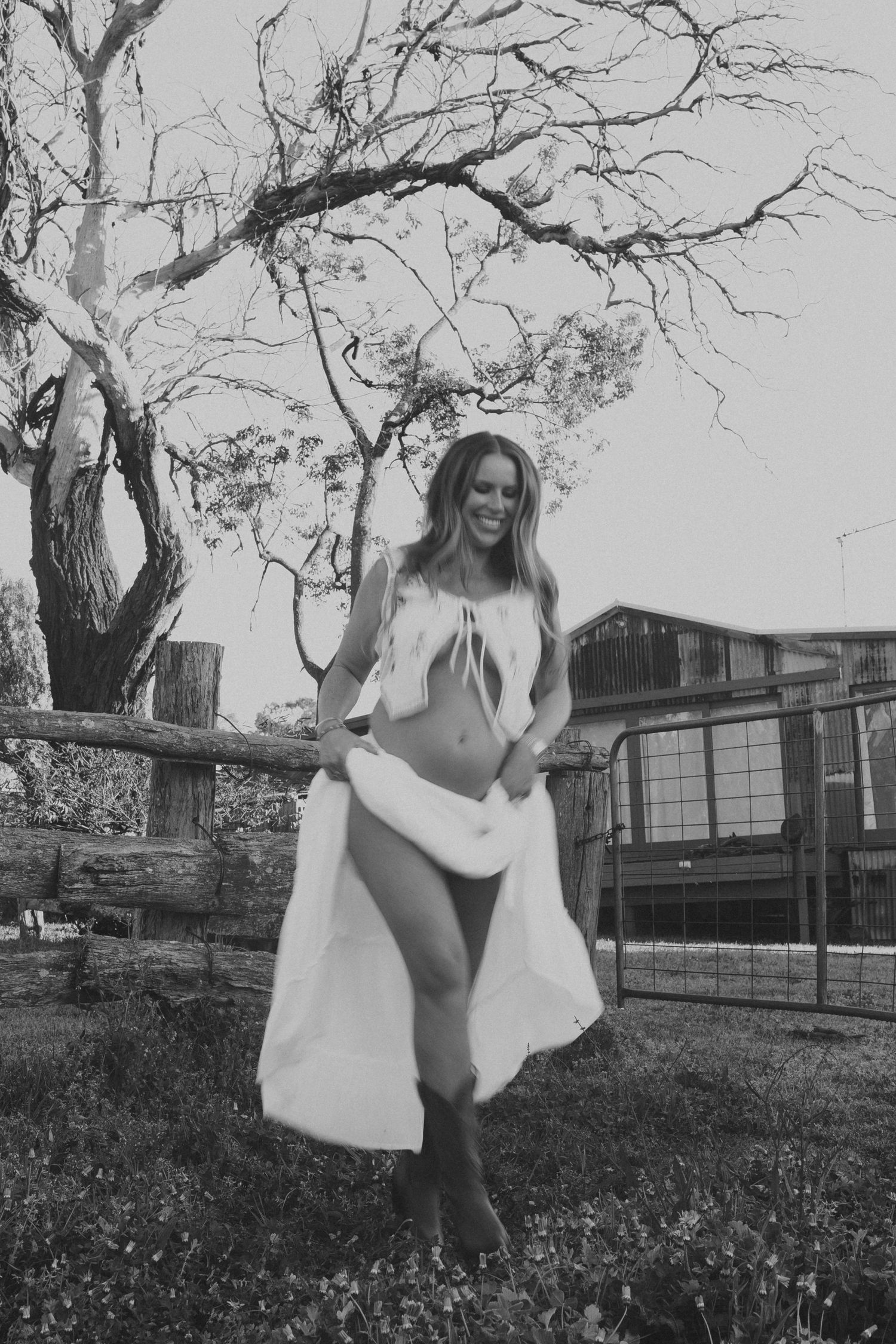 Black and white photo of smiling woman in white outfit near rustic fence and bare tree.