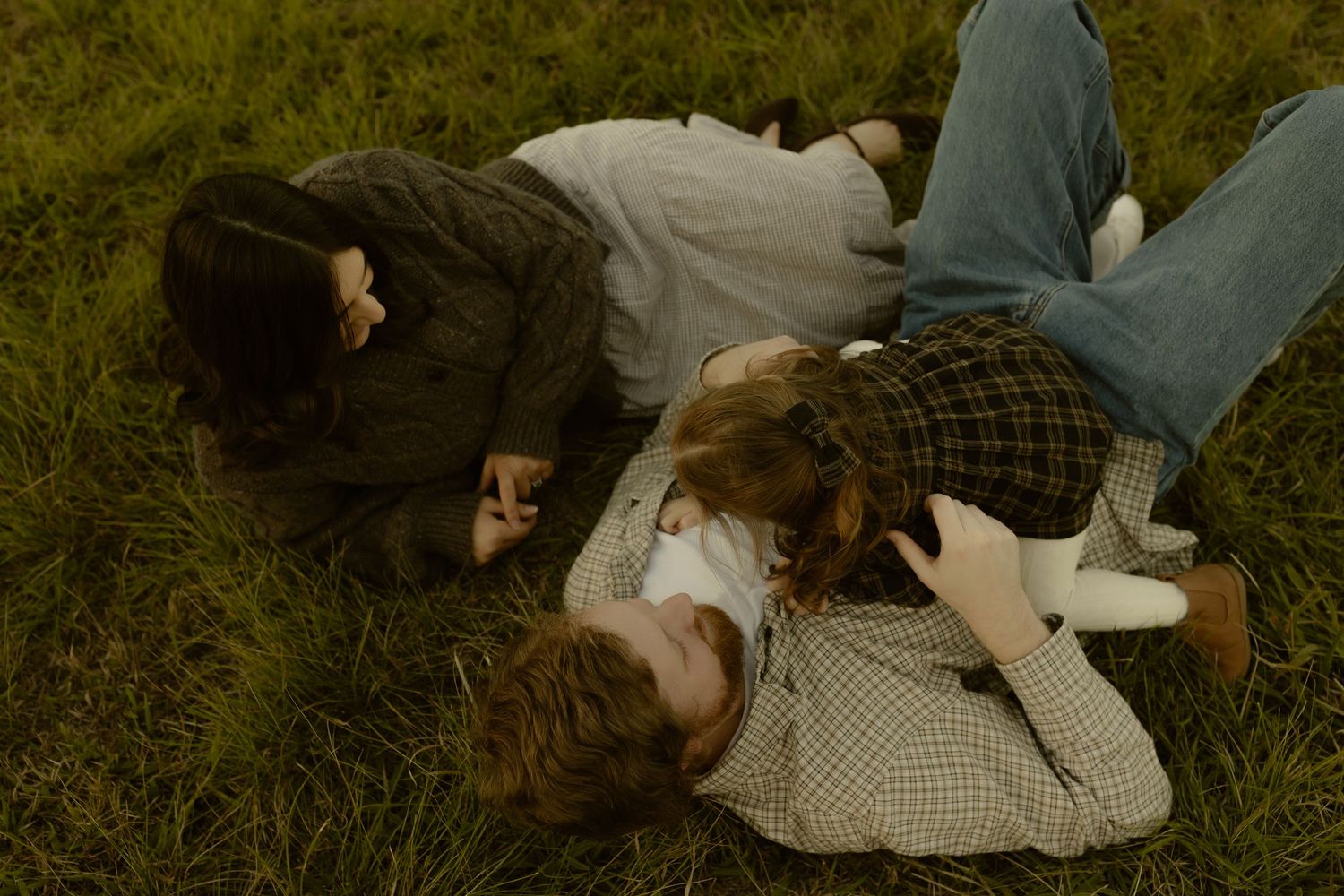 Three young people lying on green grass, laughing and playing together in a candid outdoor moment.