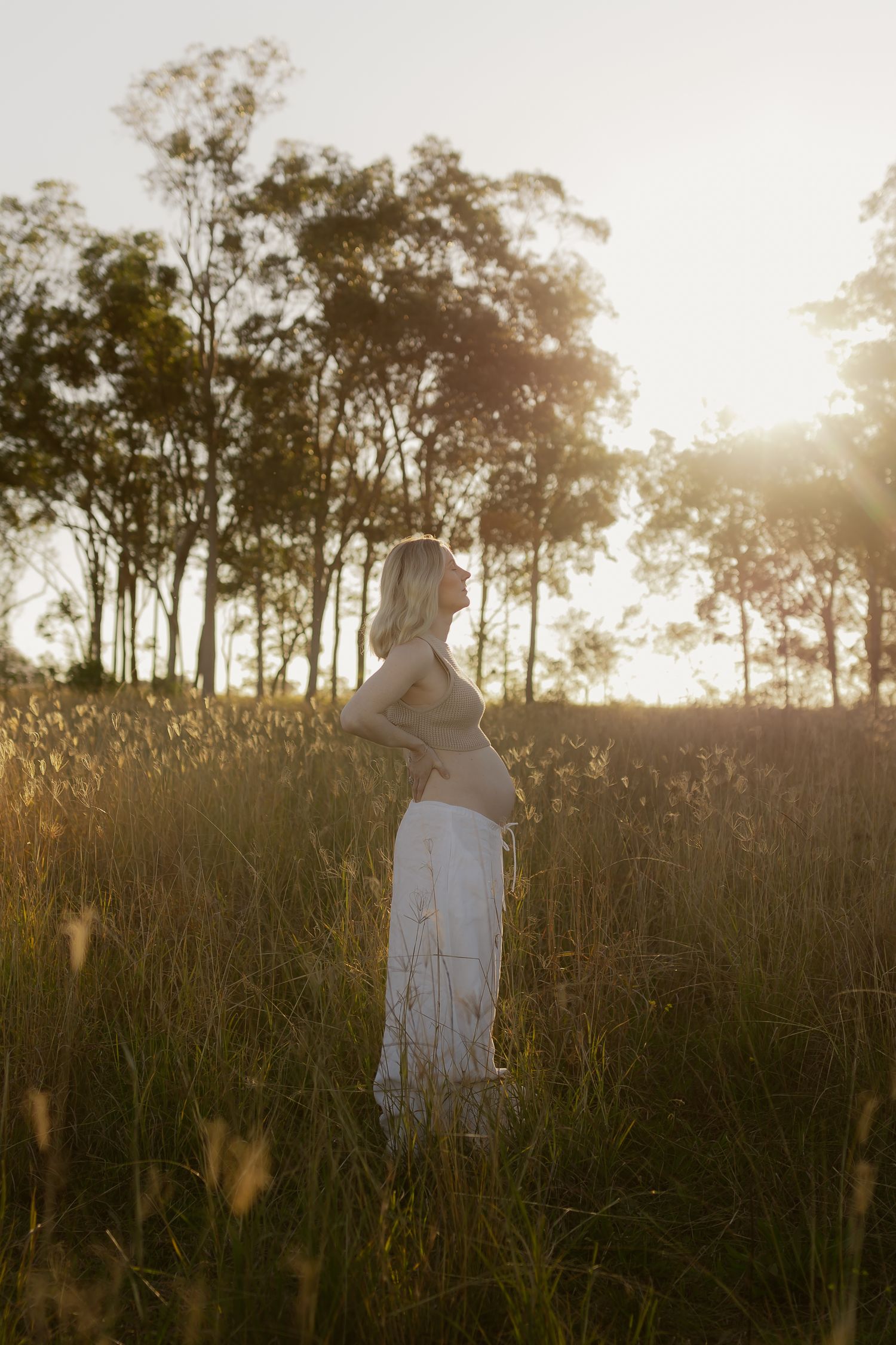 Pregnant woman in white skirt standing in golden sunset field with tall grass and trees in background.