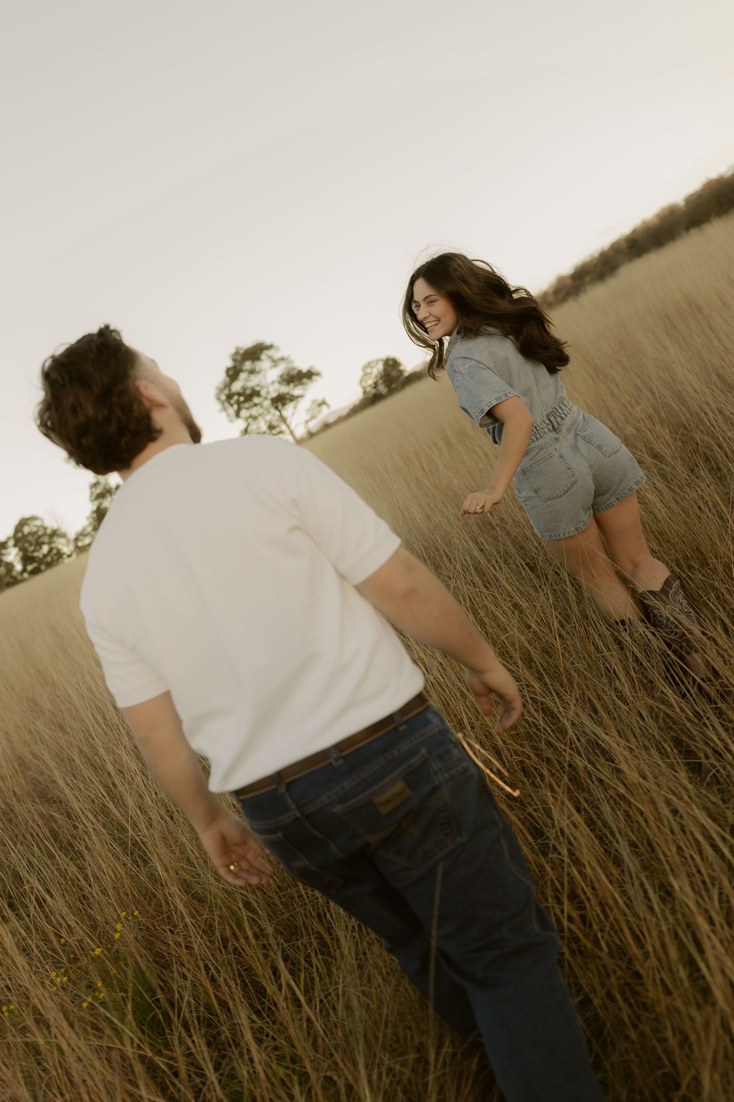 A couple walks through tall golden grass in a field at dusk, woman smiling back at man.