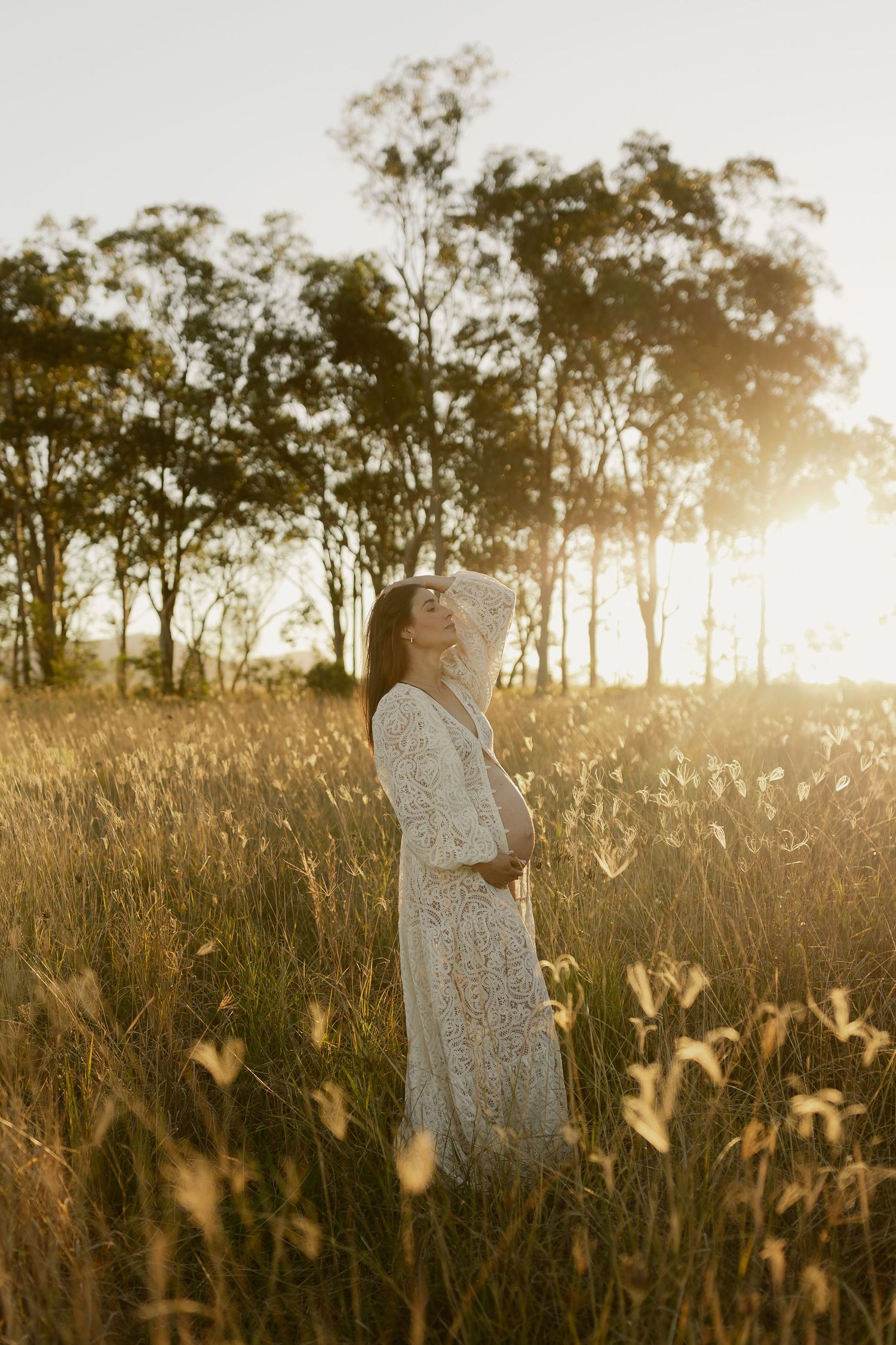 Pregnant woman in white lace dress standing in sunlit golden field with trees at sunset.