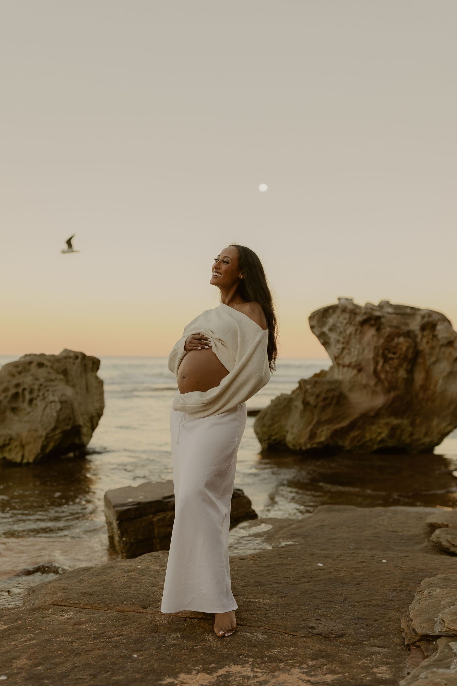 Pregnant woman in white dress cradling baby bump on rocky beach at sunset with ocean and moon backdrop.