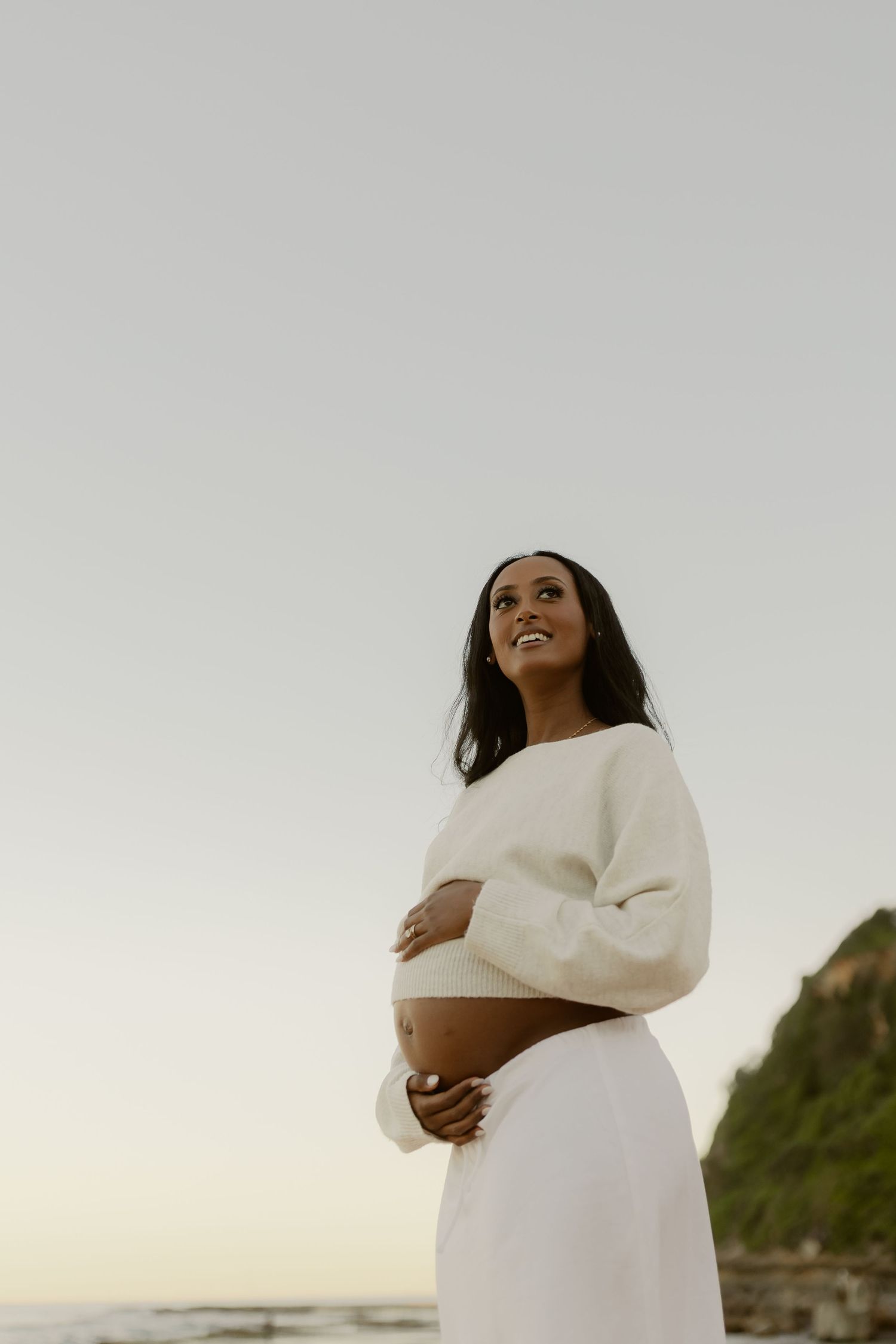 Smiling pregnant woman in white crop sweater and skirt cradling baby bump at beach sunset.