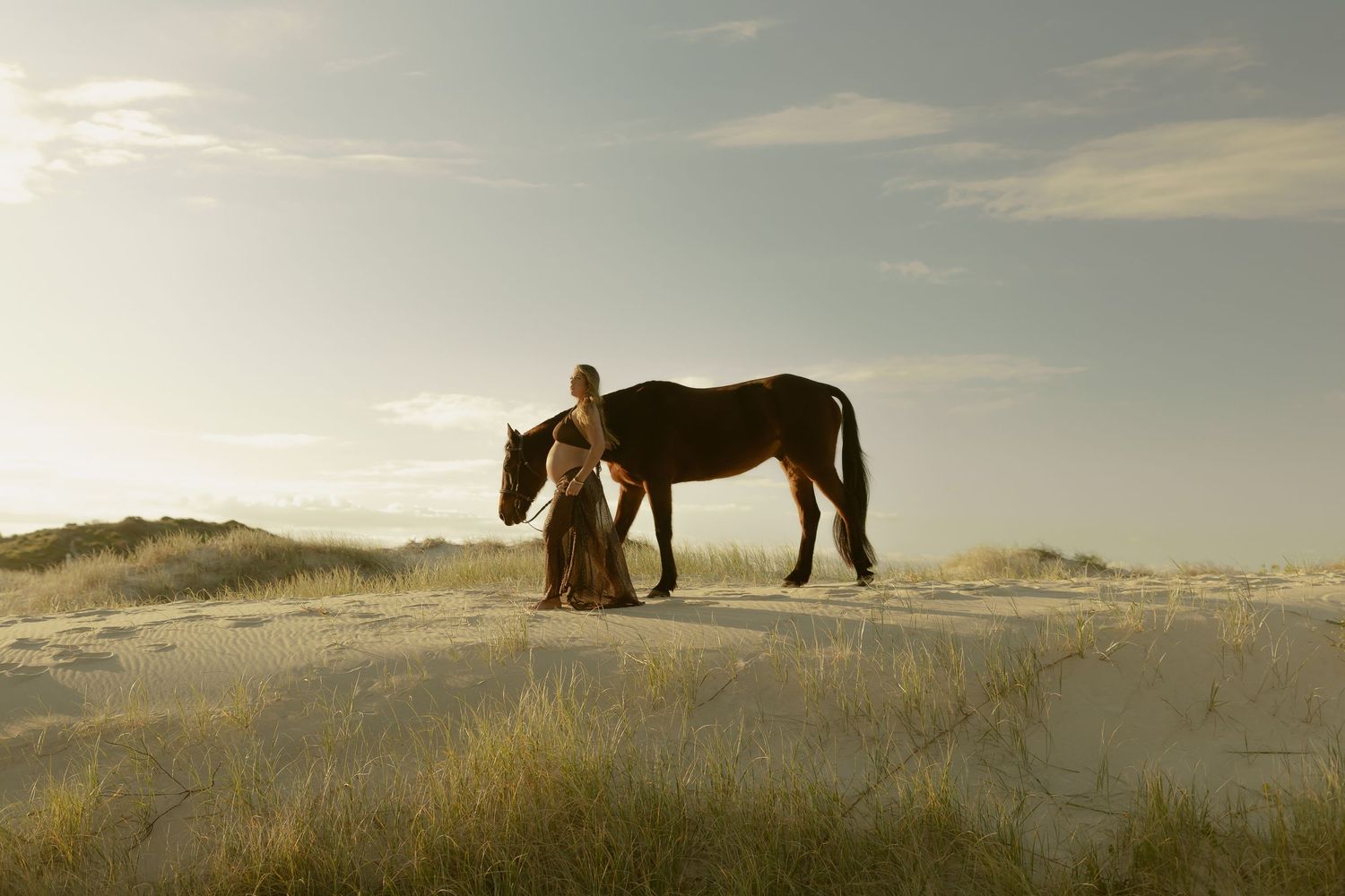 Woman in flowing dress stands beside brown horse on sunlit sand dunes with coastal backdrop.