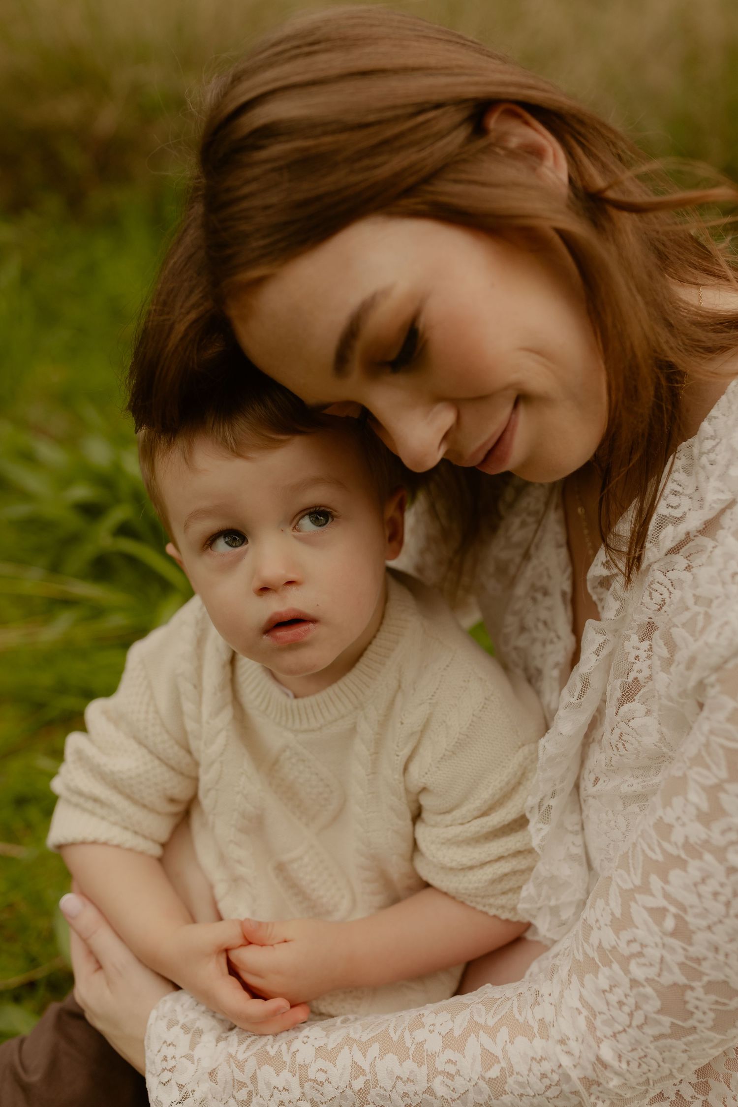 A smiling mother in a lace dress tenderly embraces her toddler boy in a cream sweater outdoors in a green field.